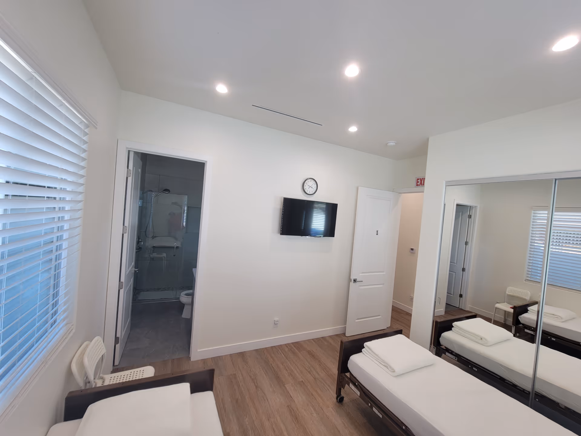 A bright shared senior living bedroom with two beds, wall TV, clock, and bathroom in doorway.