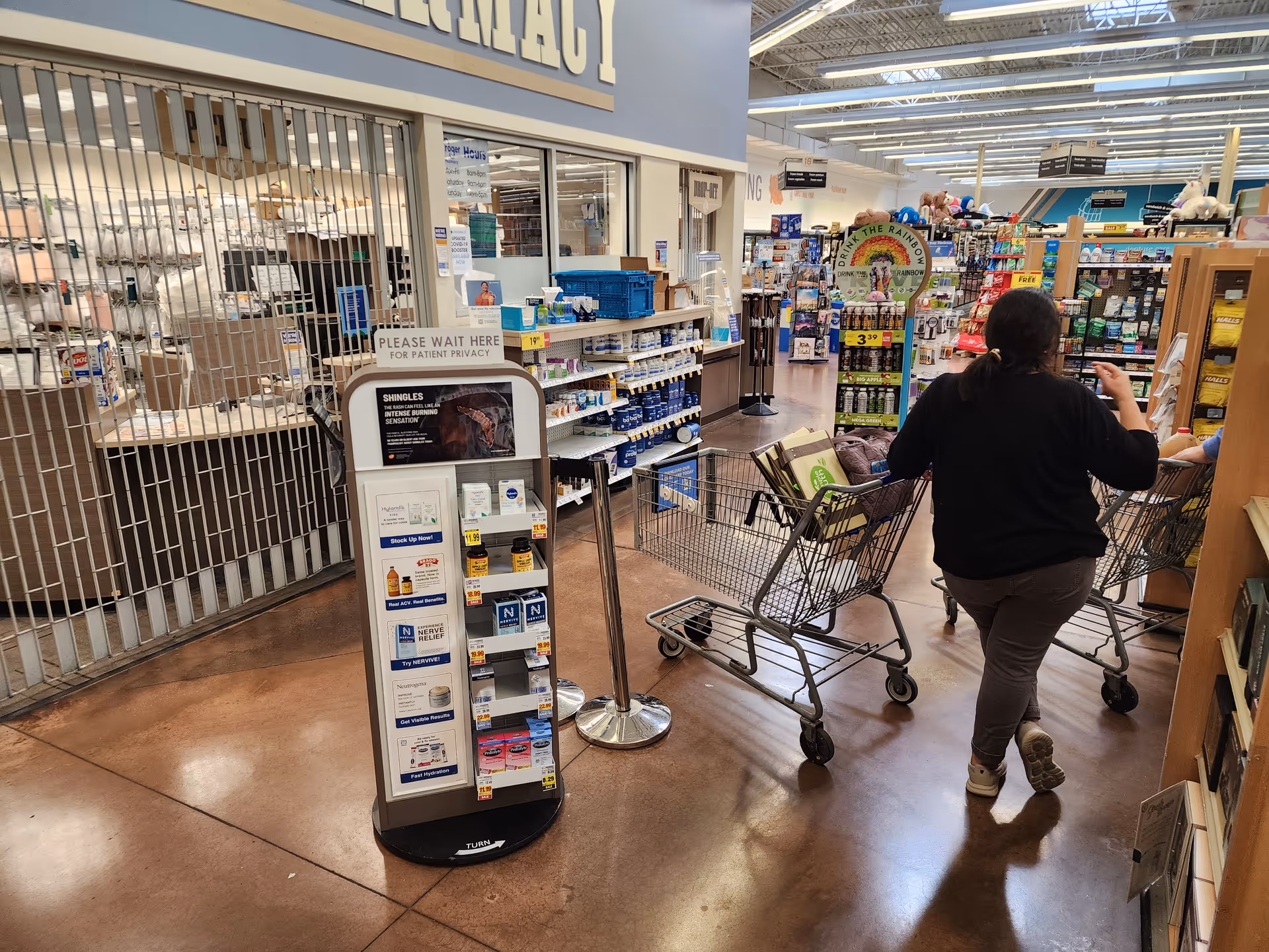 Inside a Kroger pharmacy, a shopper pushes a cart past medication aisles.