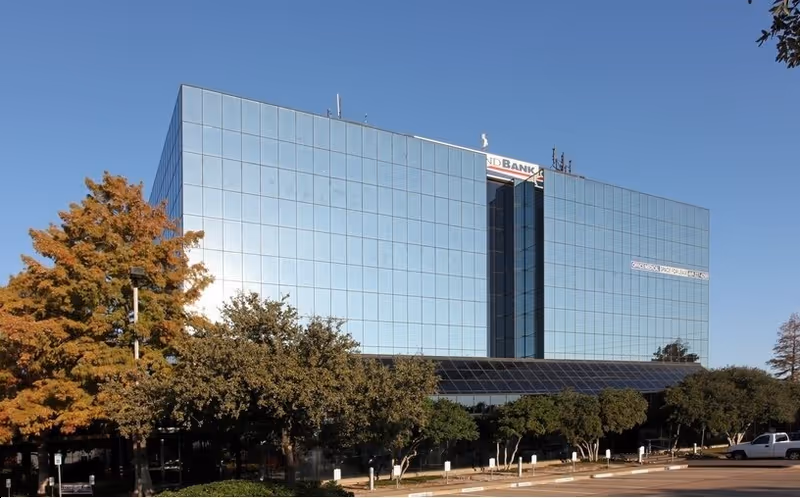 Modern glass-front office building with surrounding trees and a clear blue sky