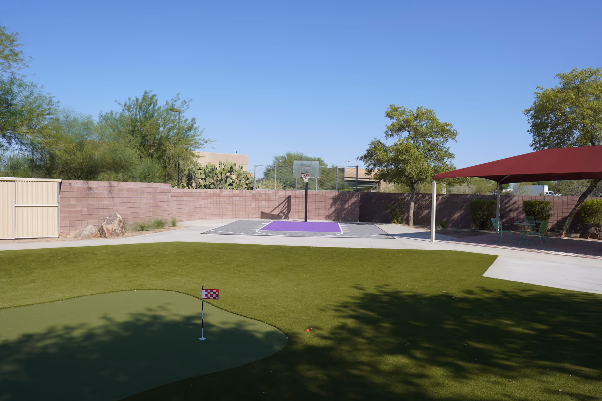 Sunny outdoor recreation area with a mini putting green, basketball hoop, and shaded seating.