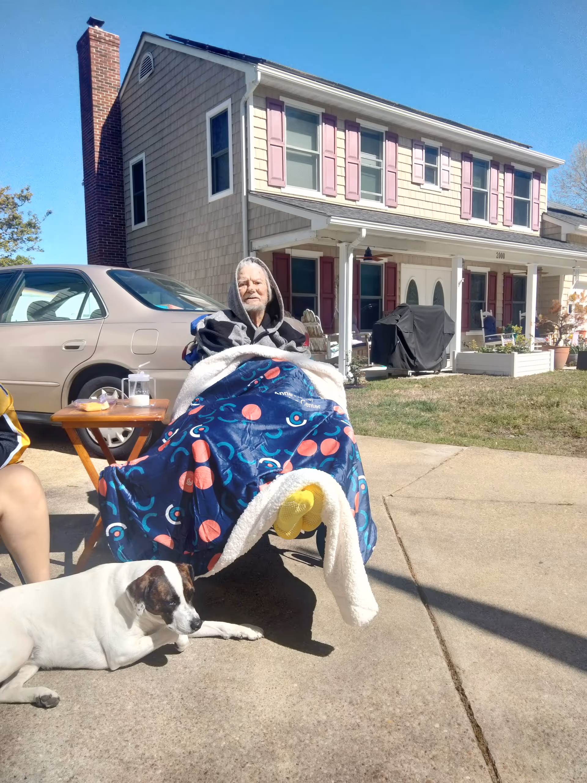 Elderly resident sits outside on a porch with a blanket, dog nearby, house in background.