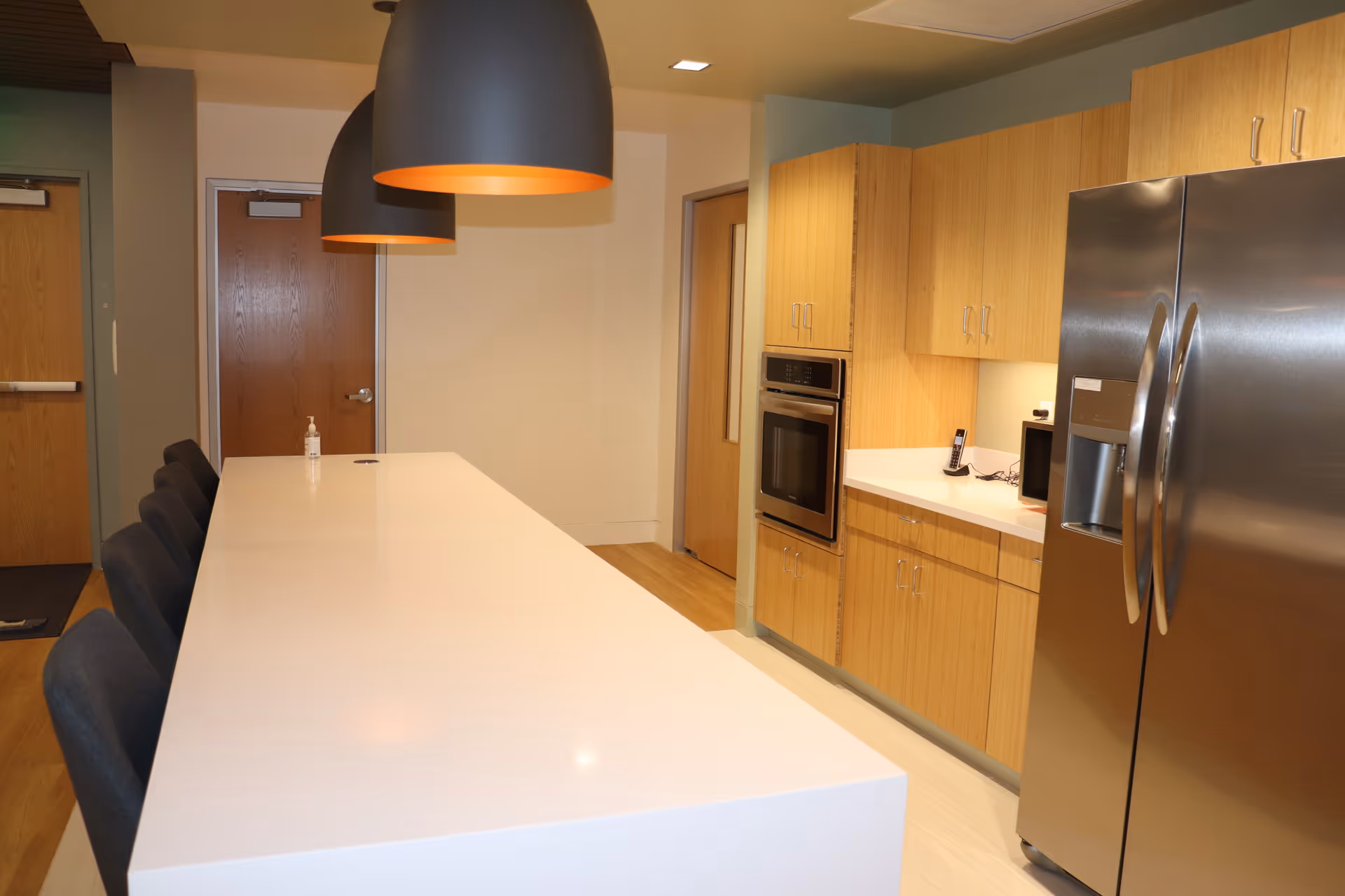 Modern communal kitchen with long white island, stainless steel fridge, and wooden cabinets.