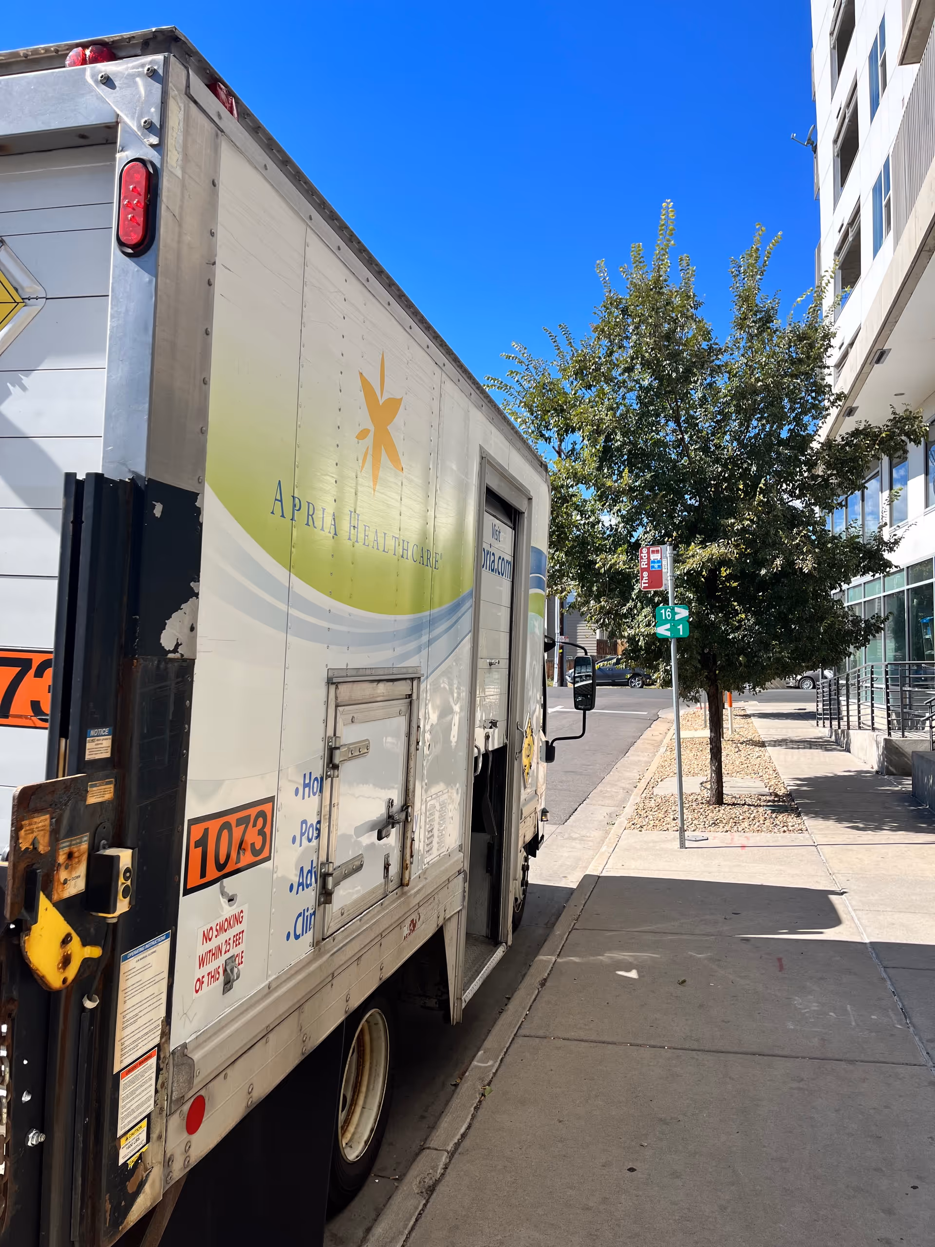 Apria Healthcare delivery truck parked along sidewalk beside a modern building and trees.