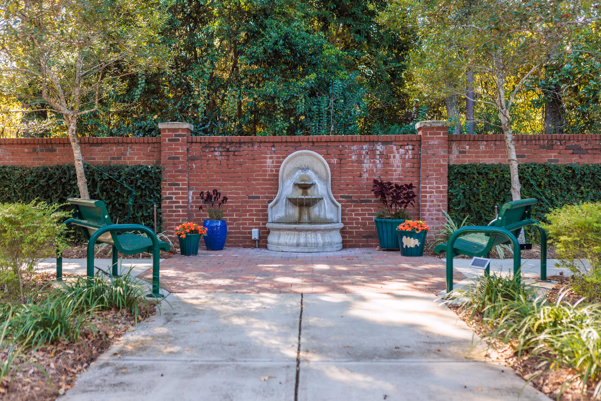 Outdoor courtyard with brick wall, central fountain, potted plants, and green benches.