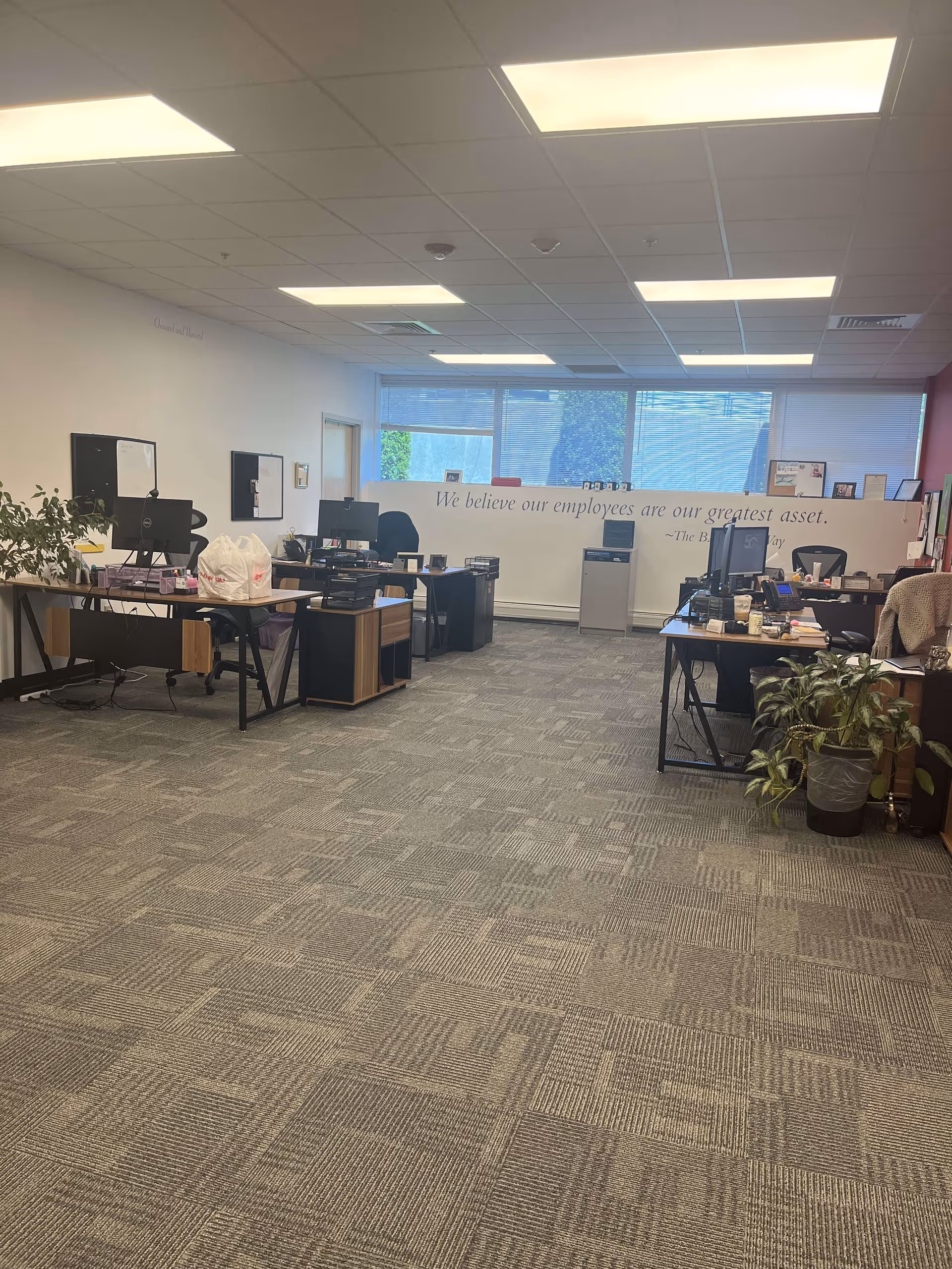 Open office area with desks, computers, plants, and a wall slogan about employees.