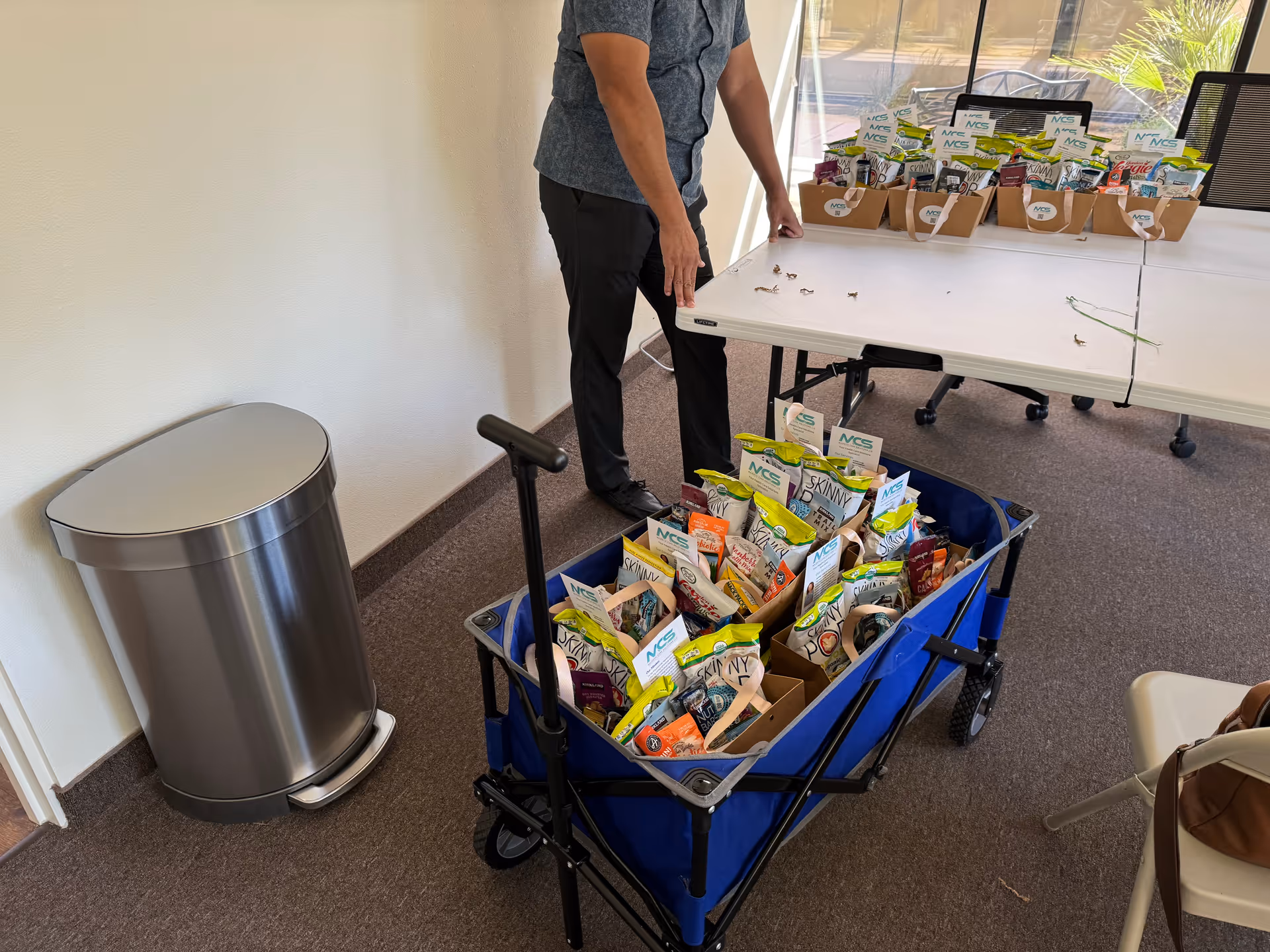 A staff member stands beside a cart filled with snack packets near a folding table.