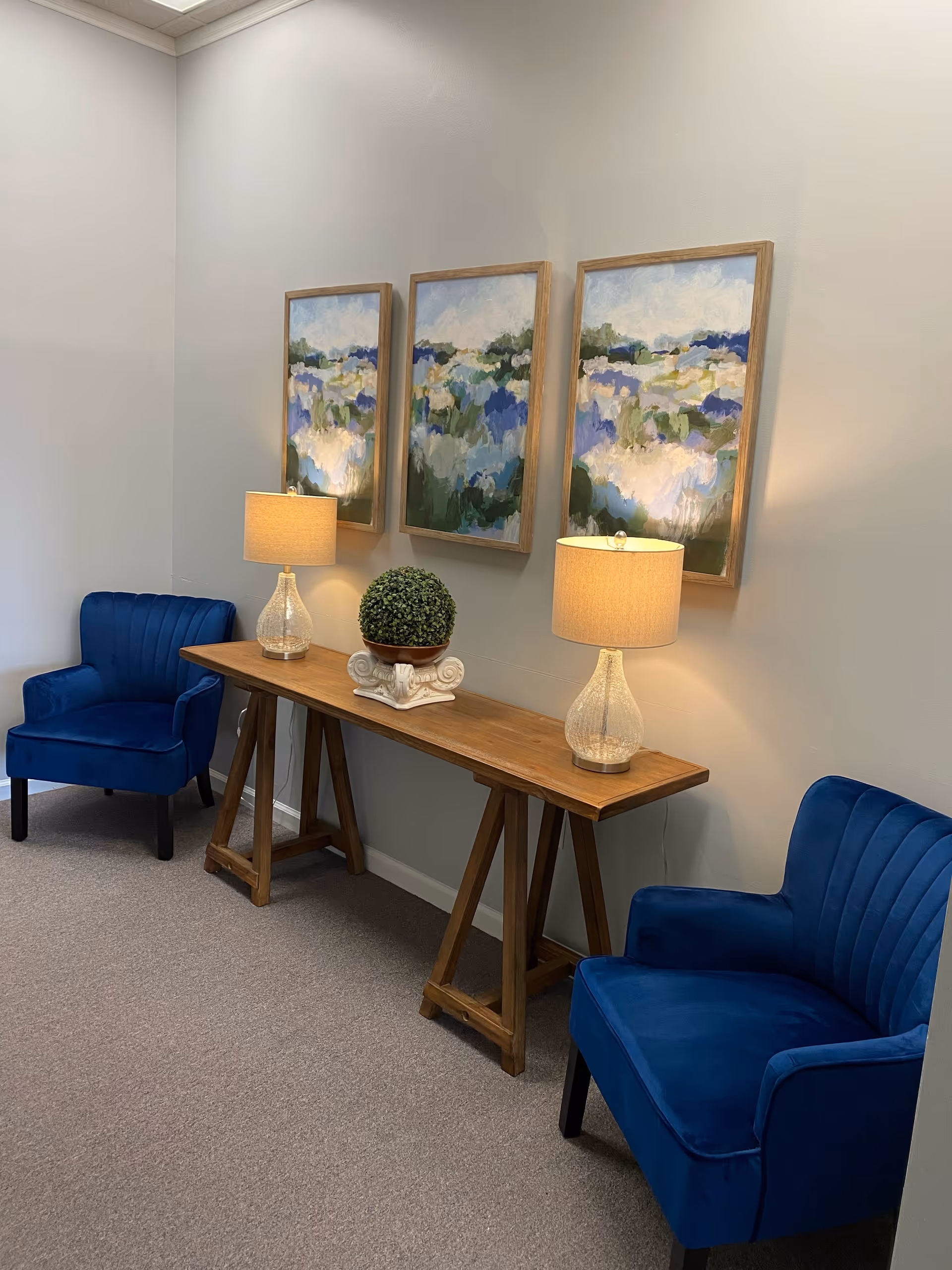 Cozy lounge with two blue chairs, a wooden console table, two lamps, and abstract paintings.