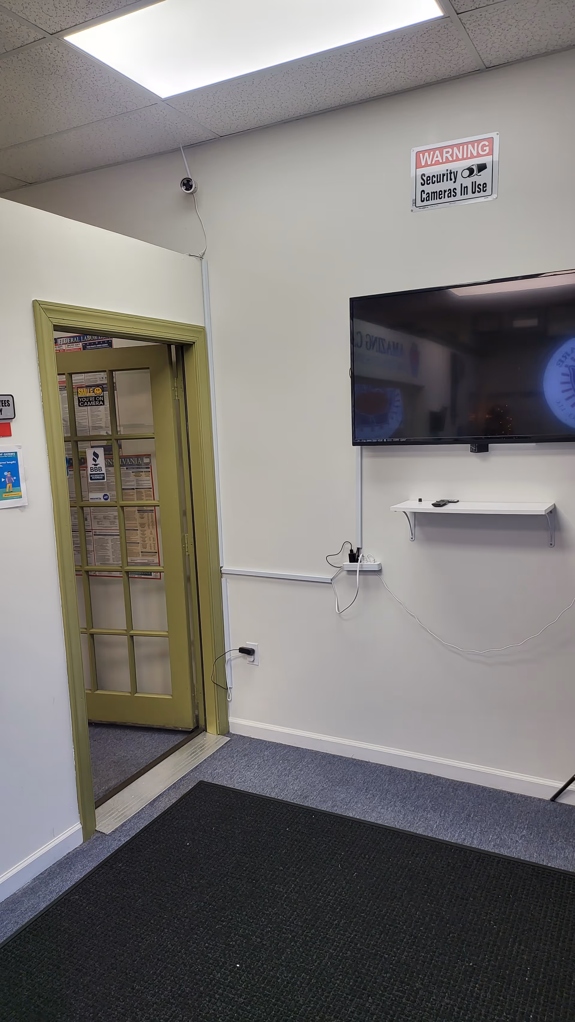 Interior hallway with security camera sign, wall-mounted TV, green door frame, and exposed cords.