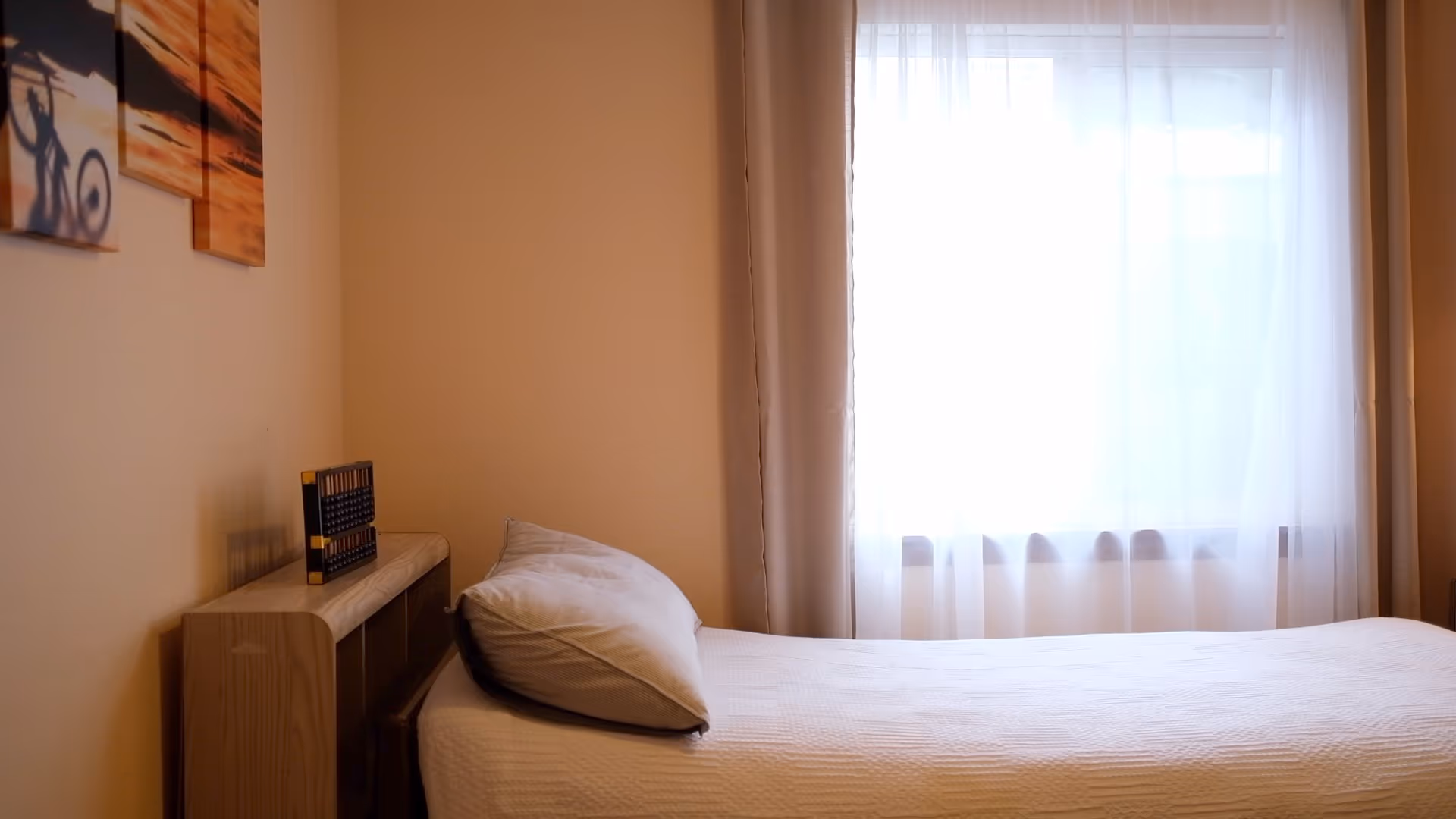A tidy bedroom features a bed, headboard shelf, and a window with sheer curtains.