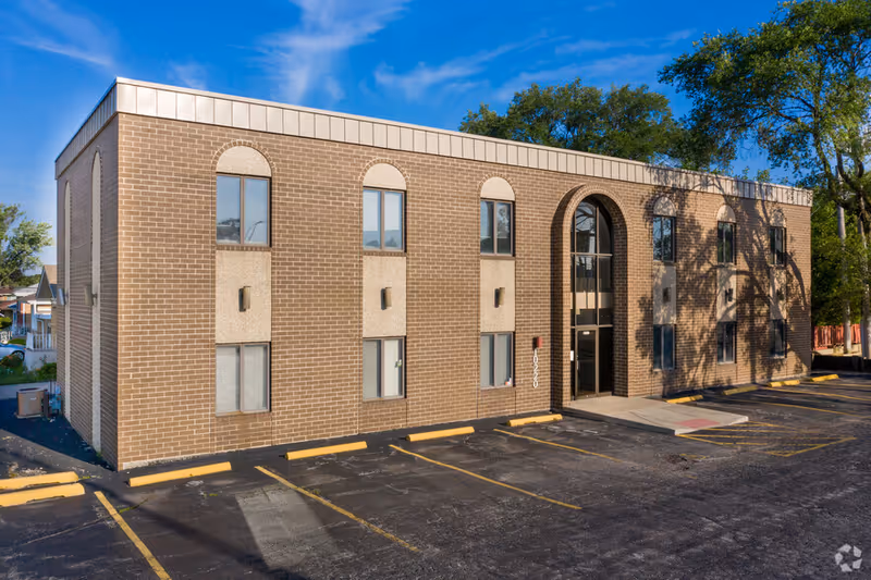 Brick two-story building with arched windows and glass entrance, adjacent parking.