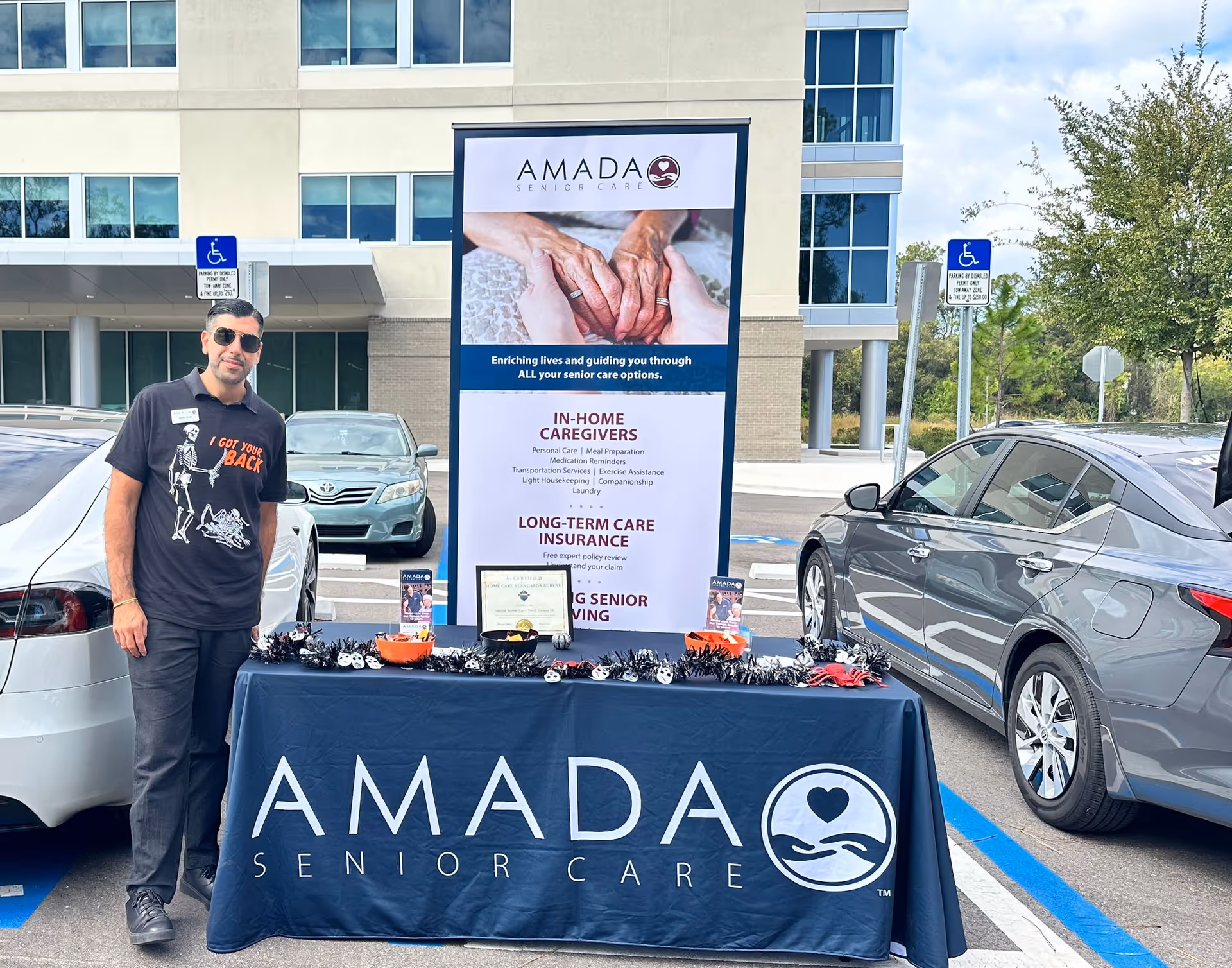 A man stands beside an Amada Senior Care promotional table outside a modern building.