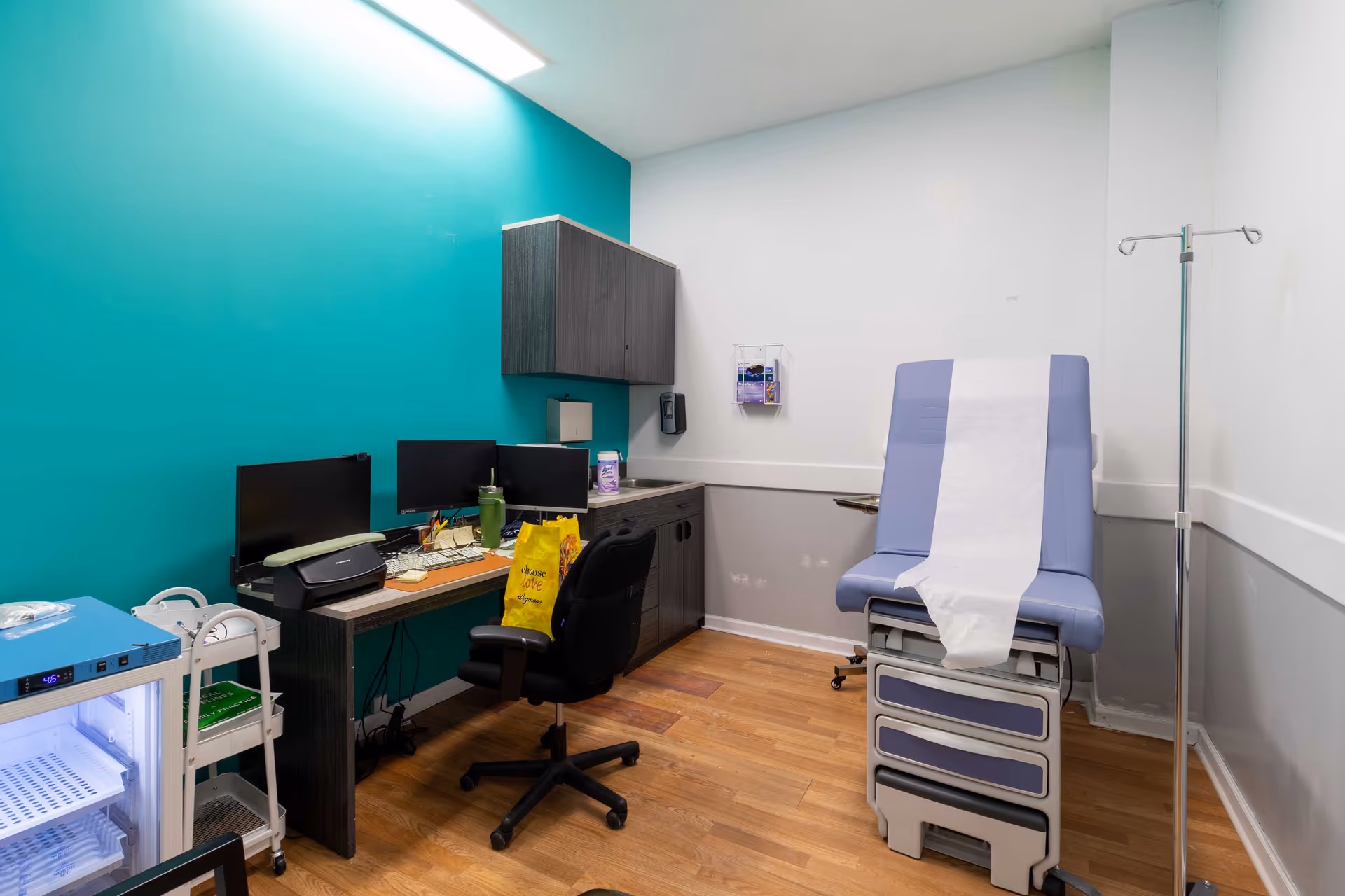 Medical examination room with teal accent wall, desk, monitors, exam chair, and IV pole.