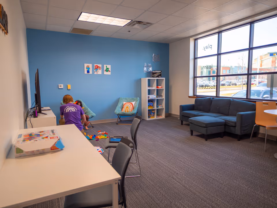 Bright playroom with blue wall, shelves, and a blue couch; residents play near the window.