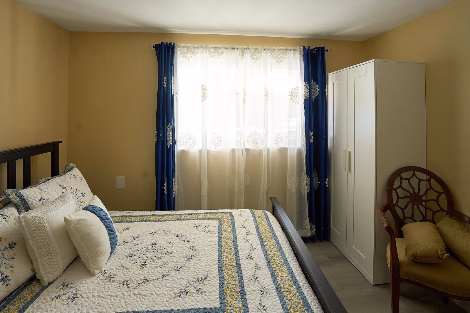 A cozy bedroom with a quilted bed, window curtains, a white wardrobe, and a chair.