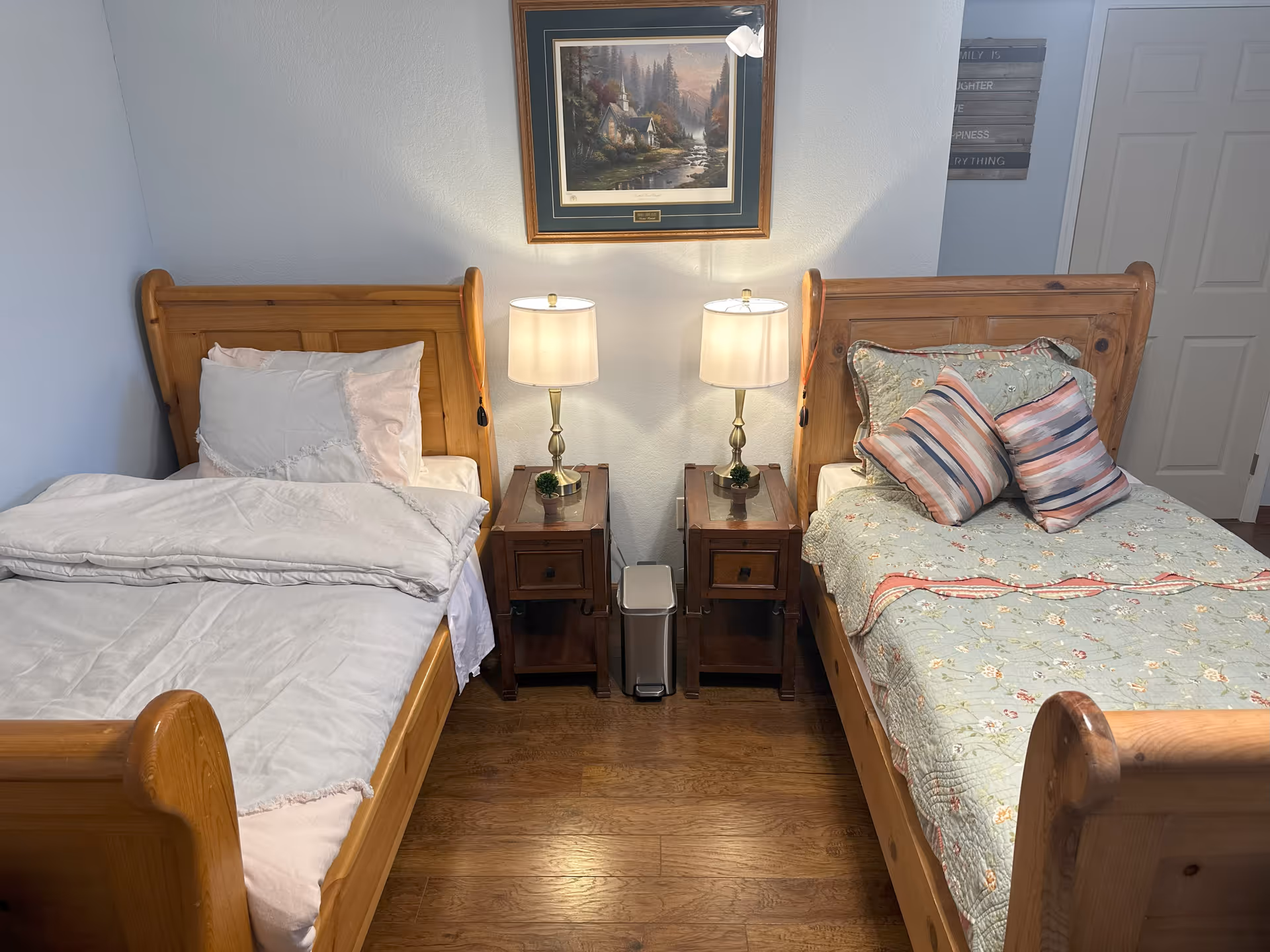 Two wooden twin beds with nightstands, lamps, and a framed landscape painting in a shared bedroom.