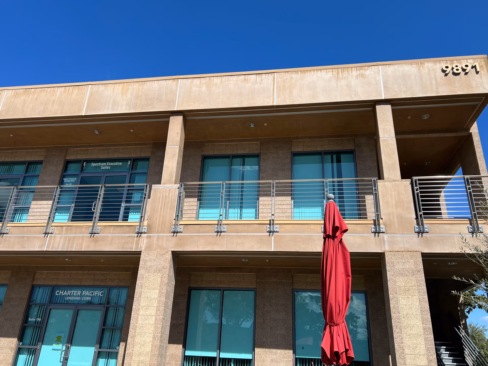 Two-story tan building exterior with glass doors, balcony railings, and a red umbrella.