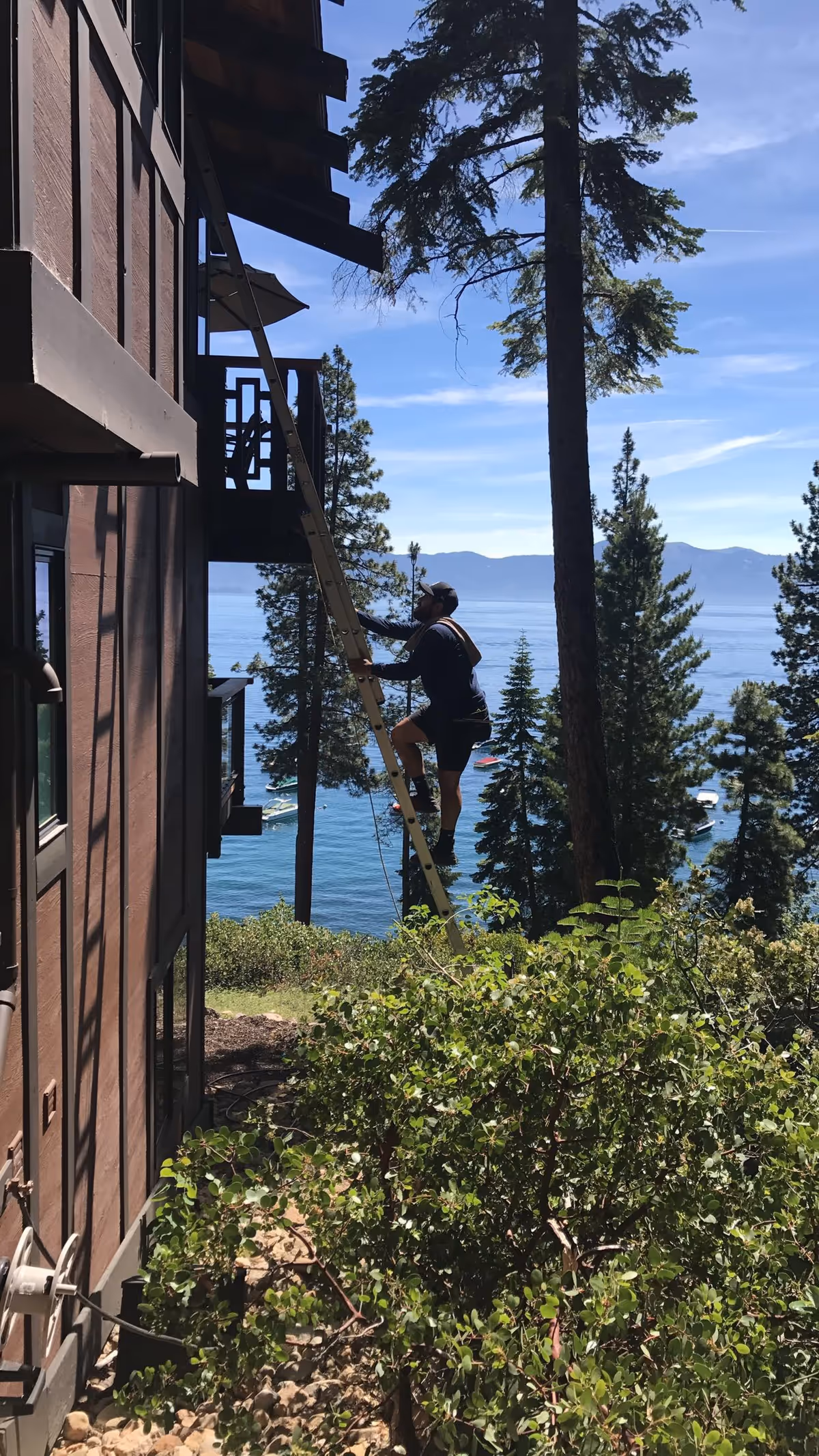 A window cleaner climbs a ladder beside a brown building with a scenic lake view.