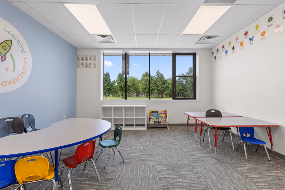 Bright classroom-style activity room with tables, colorful chairs, shelves, and a large window.
