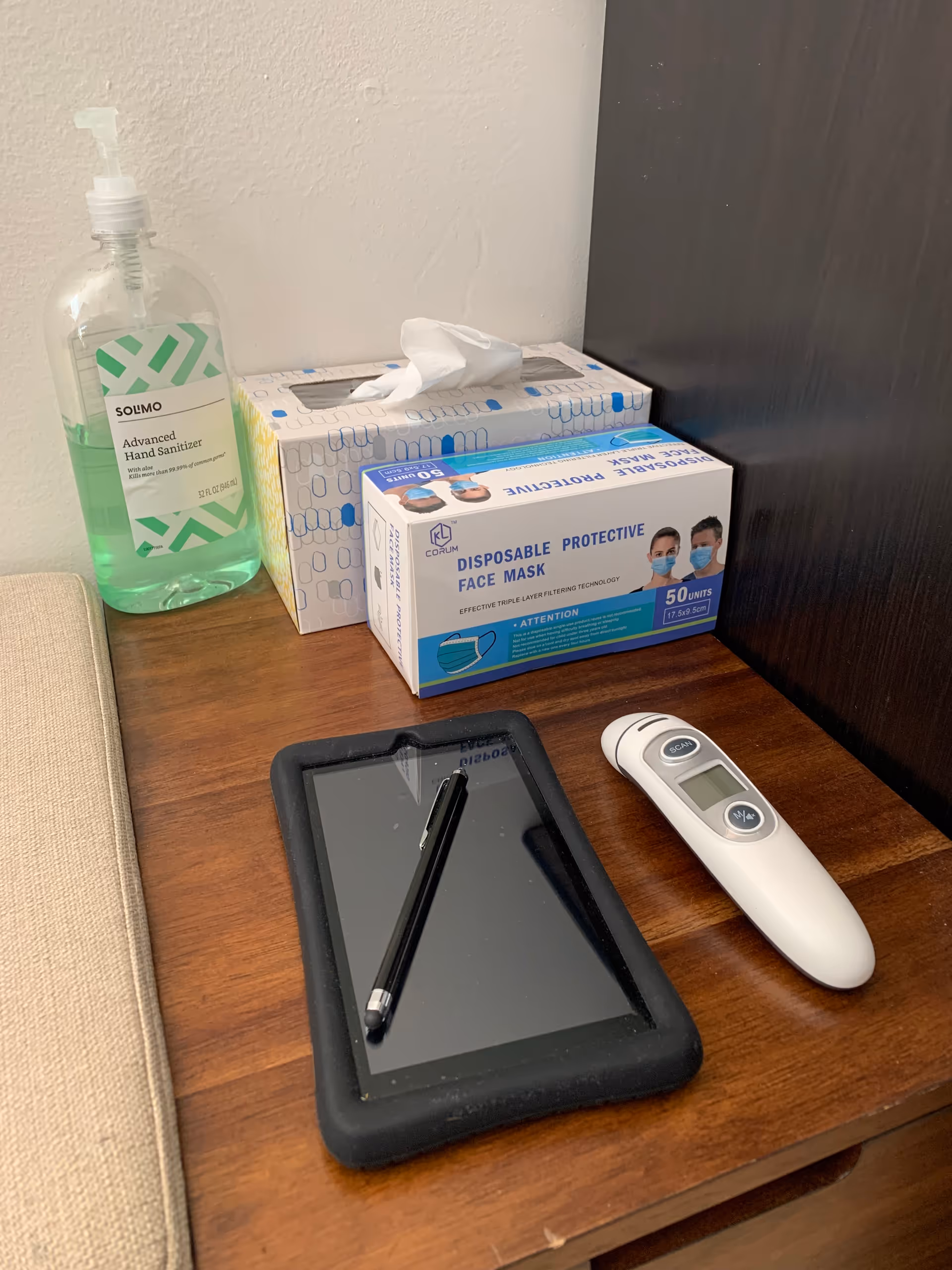 A wooden table holds hand sanitizer, tissues, a mask box, a tablet, and a digital thermometer.