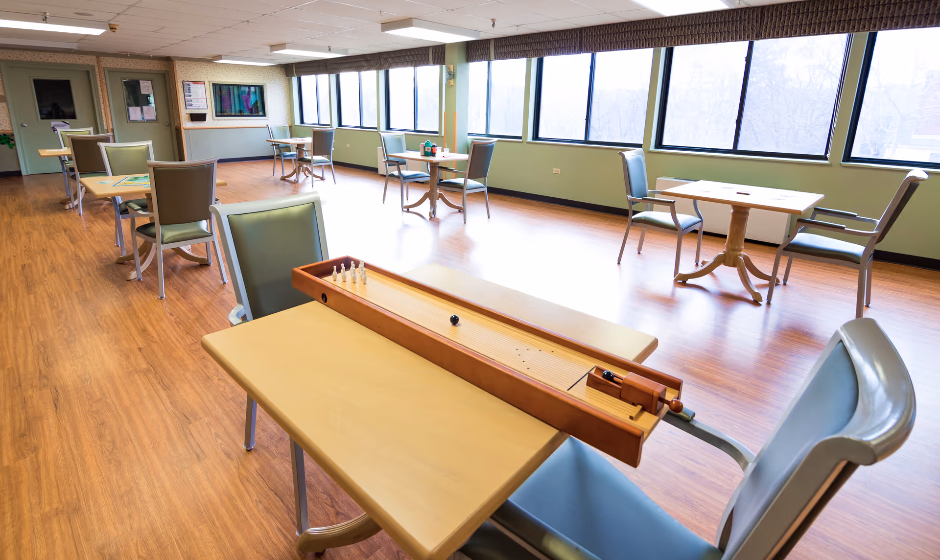 Bright communal recreation room with multiple tables, chairs, and a shuffleboard game.