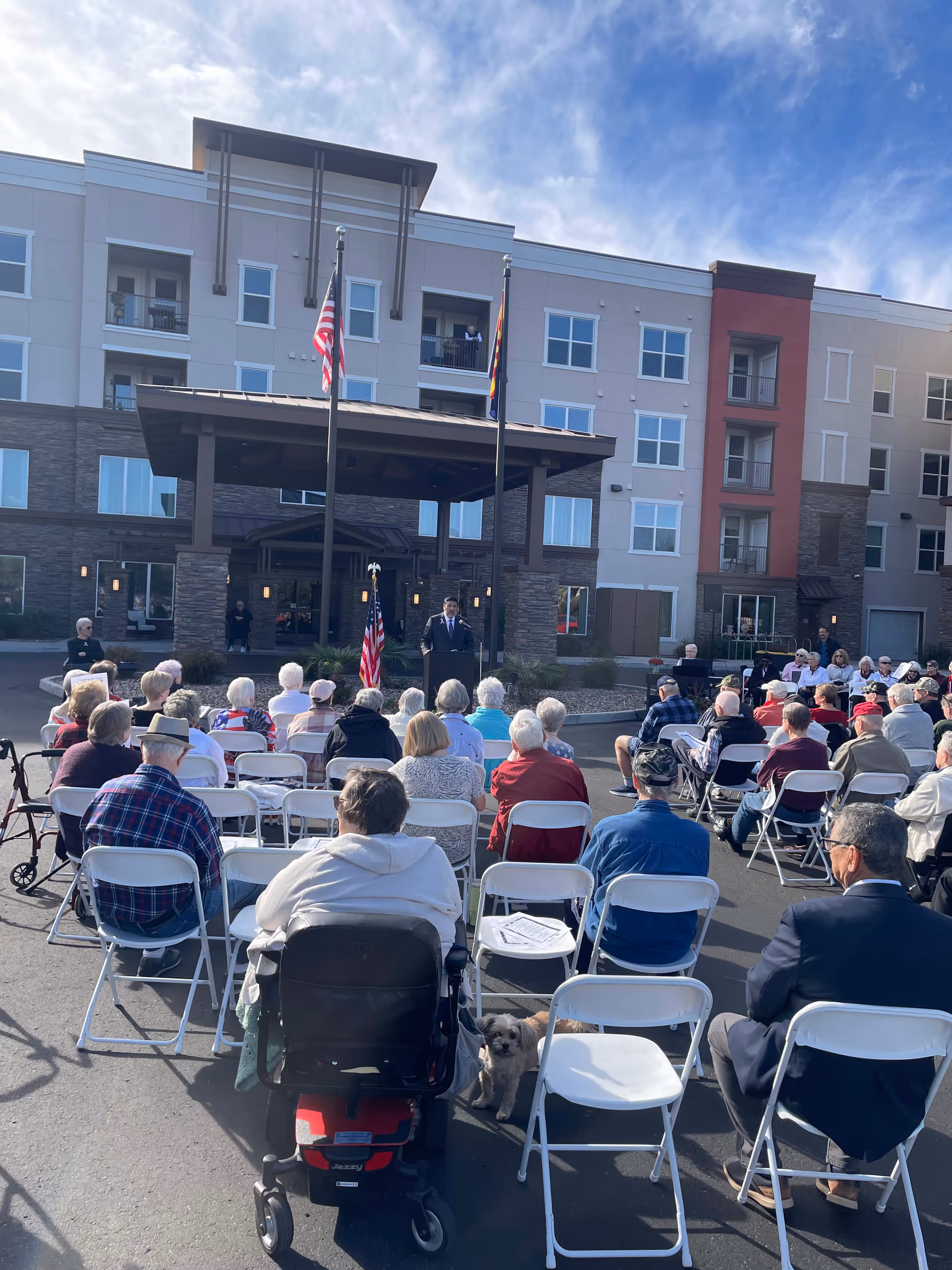Senior residents seated outdoors in chairs, listening to a speaker with flags and the building behind.