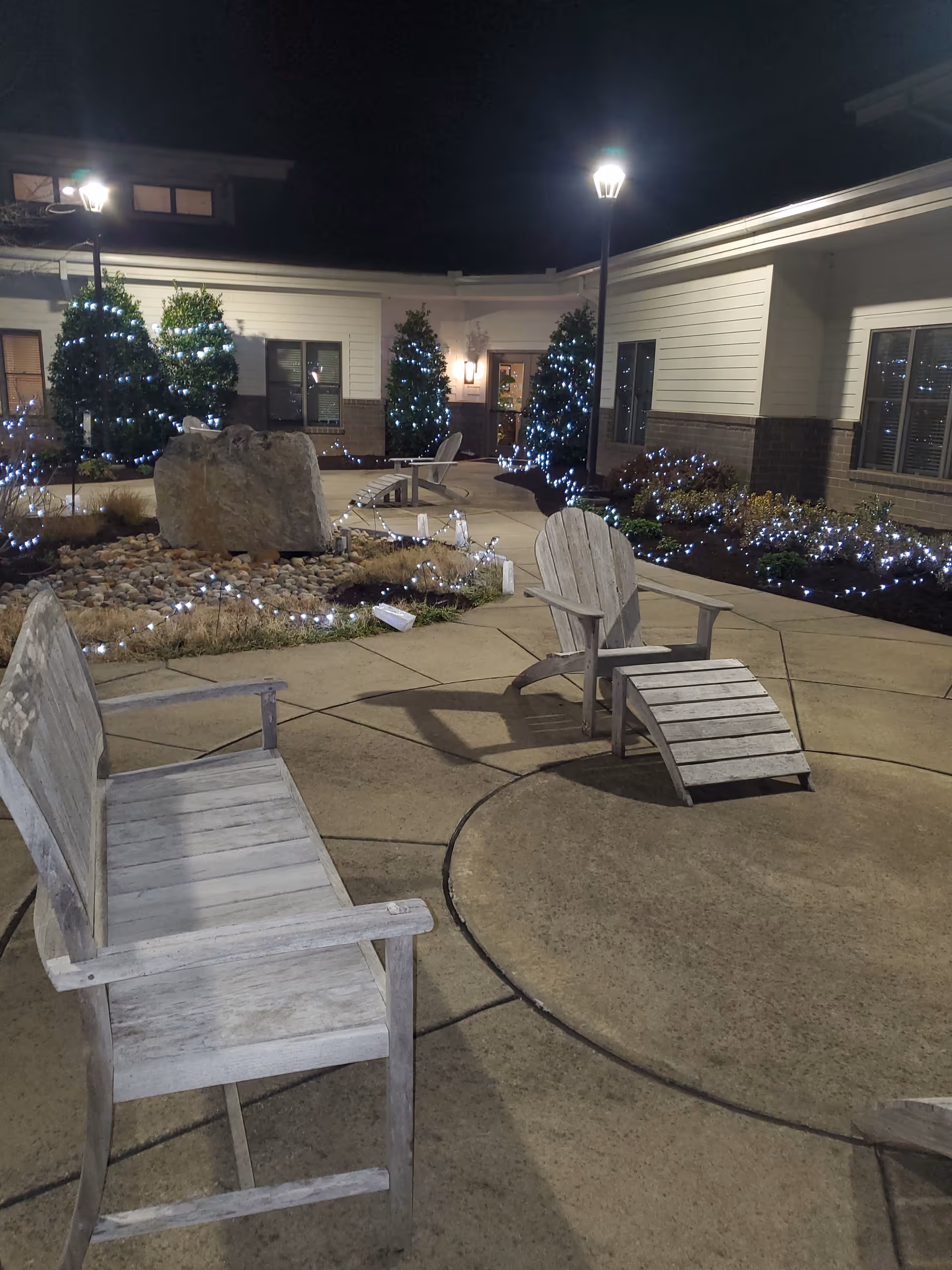 Outdoor courtyard at night with lit string lights, wooden chairs, and low shrubs.