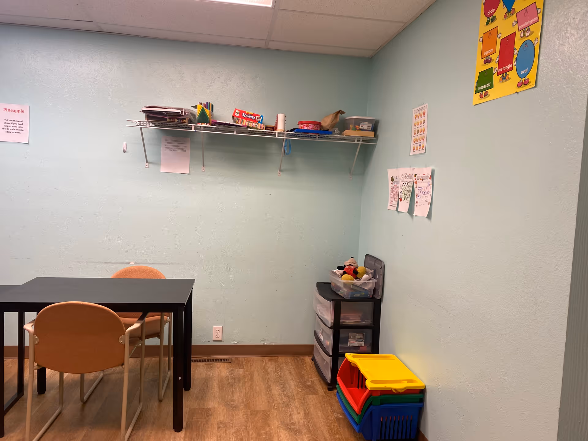 Small classroom-style activity room with table, chairs, shelves, and colorful plastic bins.