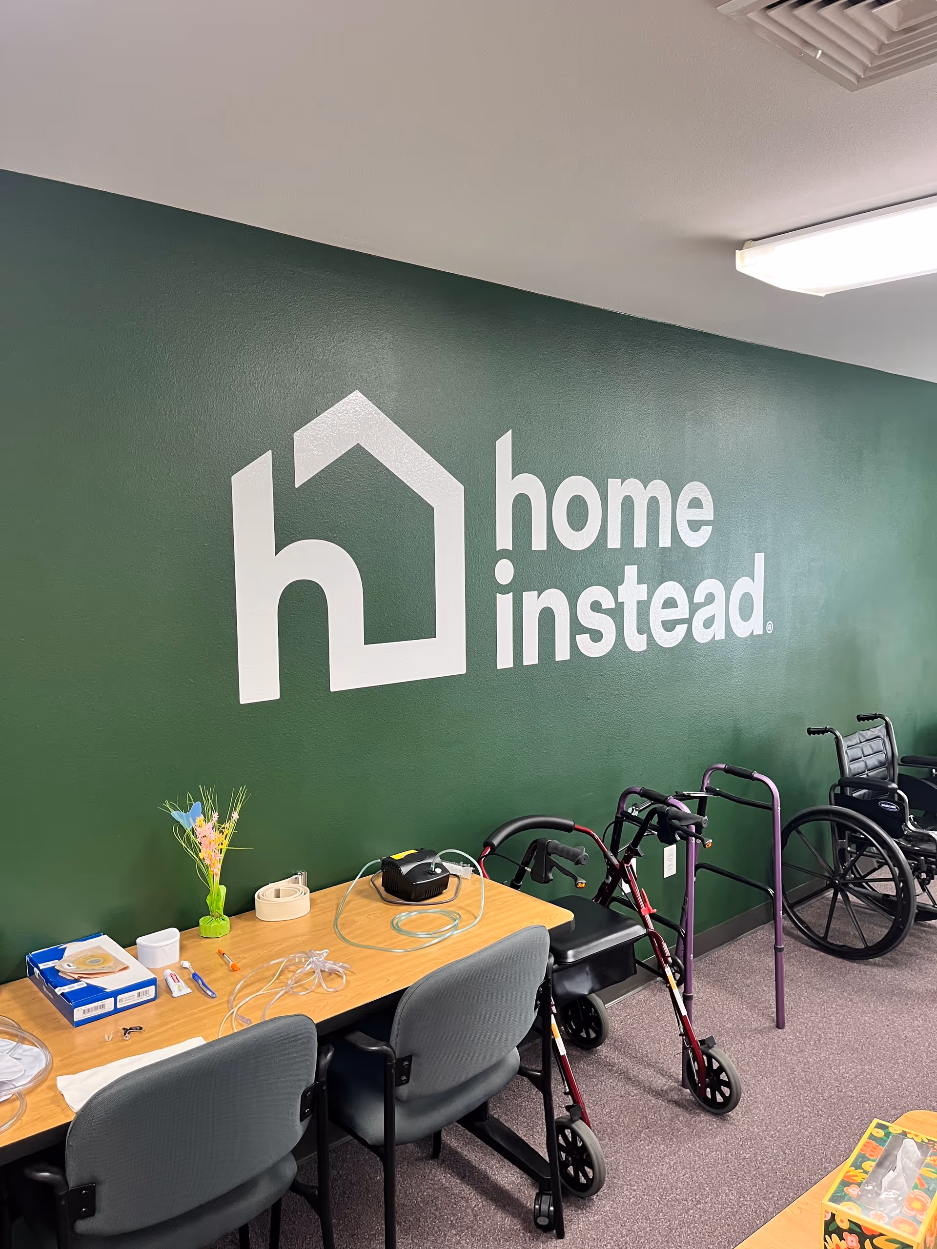 Green-walled interior shows Home Instead logo with a desk, chairs, wheelchairs, and walkers.