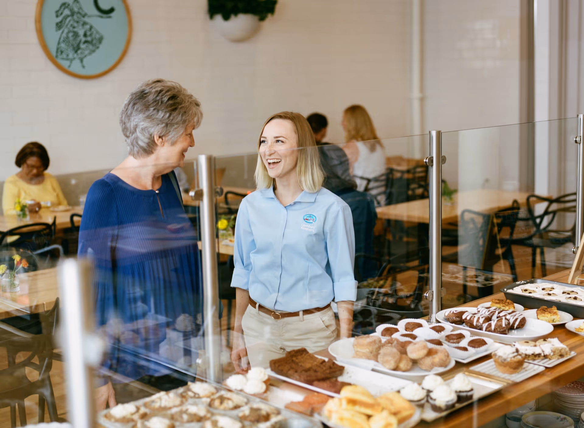 Staff member chats with a resident near a pastry display in a bright dining area.