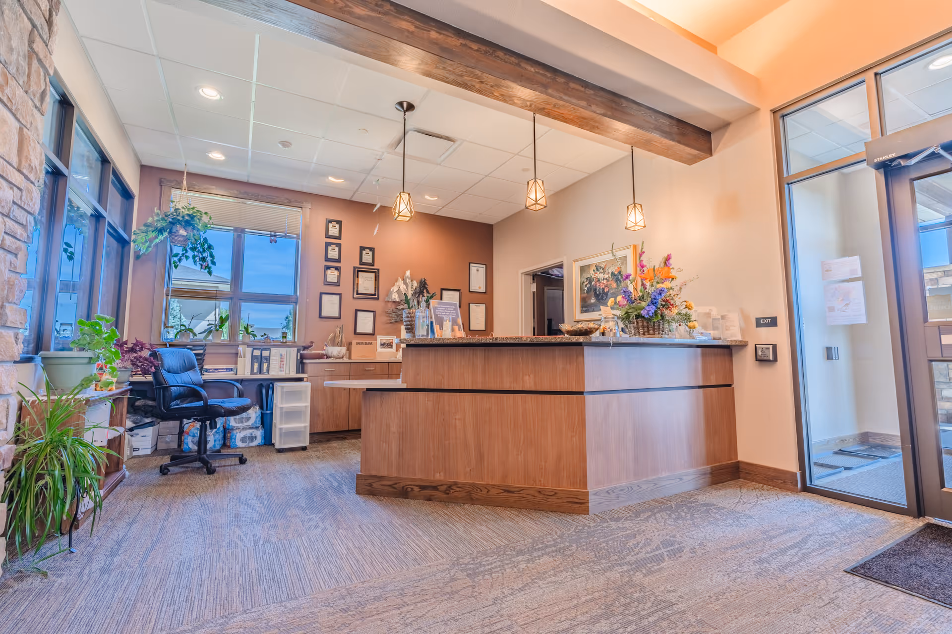 Reception desk in a warm lobby with plants, certificates, and a floral arrangement.