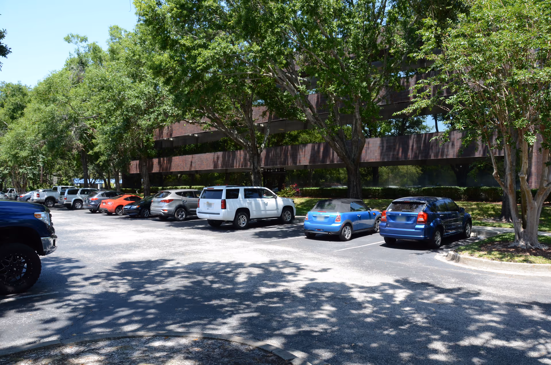Parking lot with several cars in front of a brick building shaded by trees.