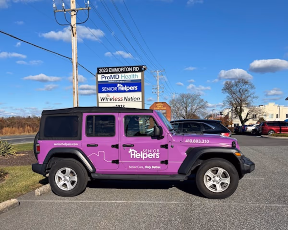 Purple Senior Helpers van parked in a lot with a facility sign behind.