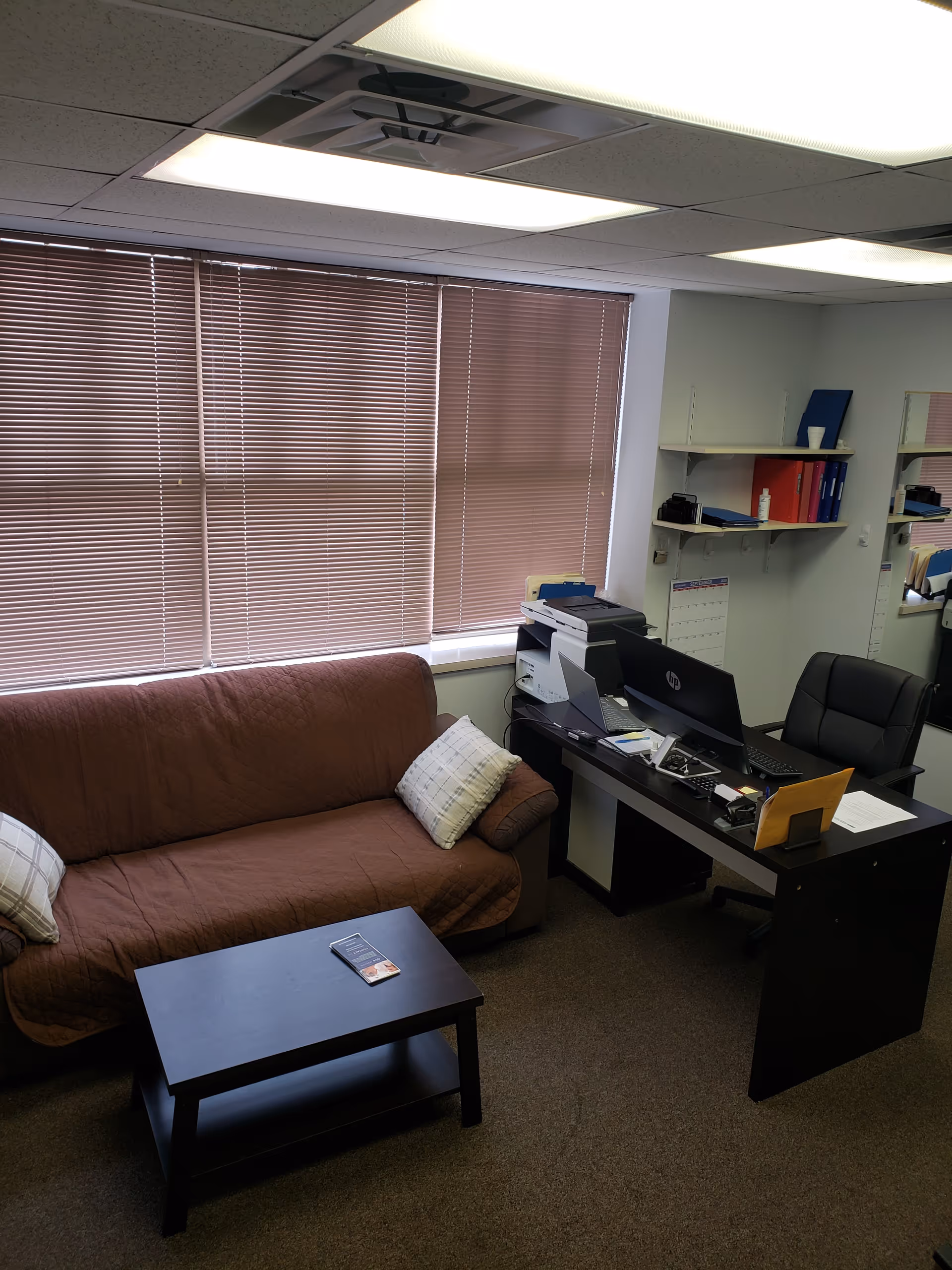 Office-like lounge area with brown sofa, coffee table, desk, computer, and blinds.