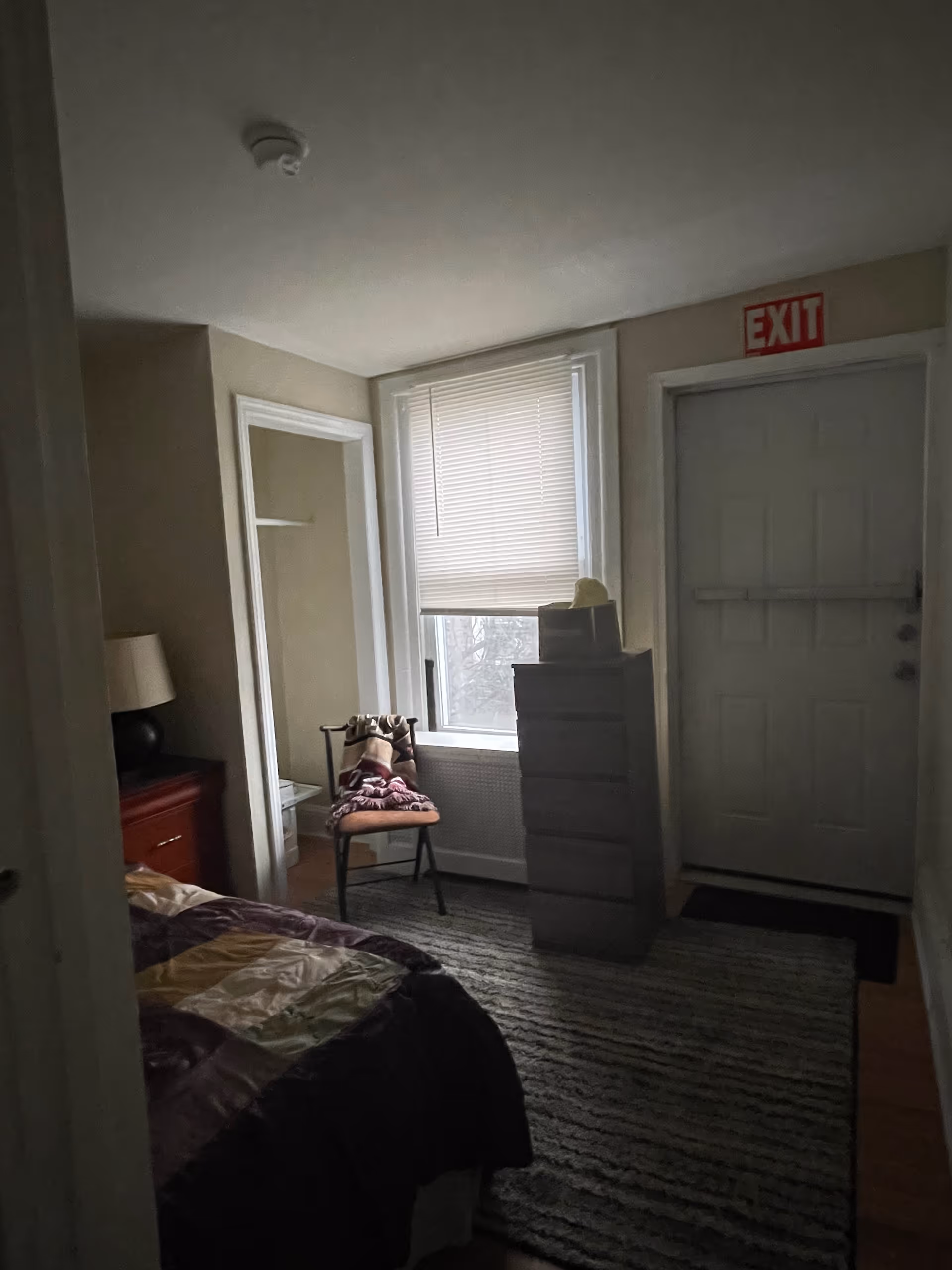 Dim bedroom with bed, dresser, window, chair, and exit door in a small apartment.
