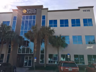 Front-view of a modern, multi-story building with palm trees and parked cars.