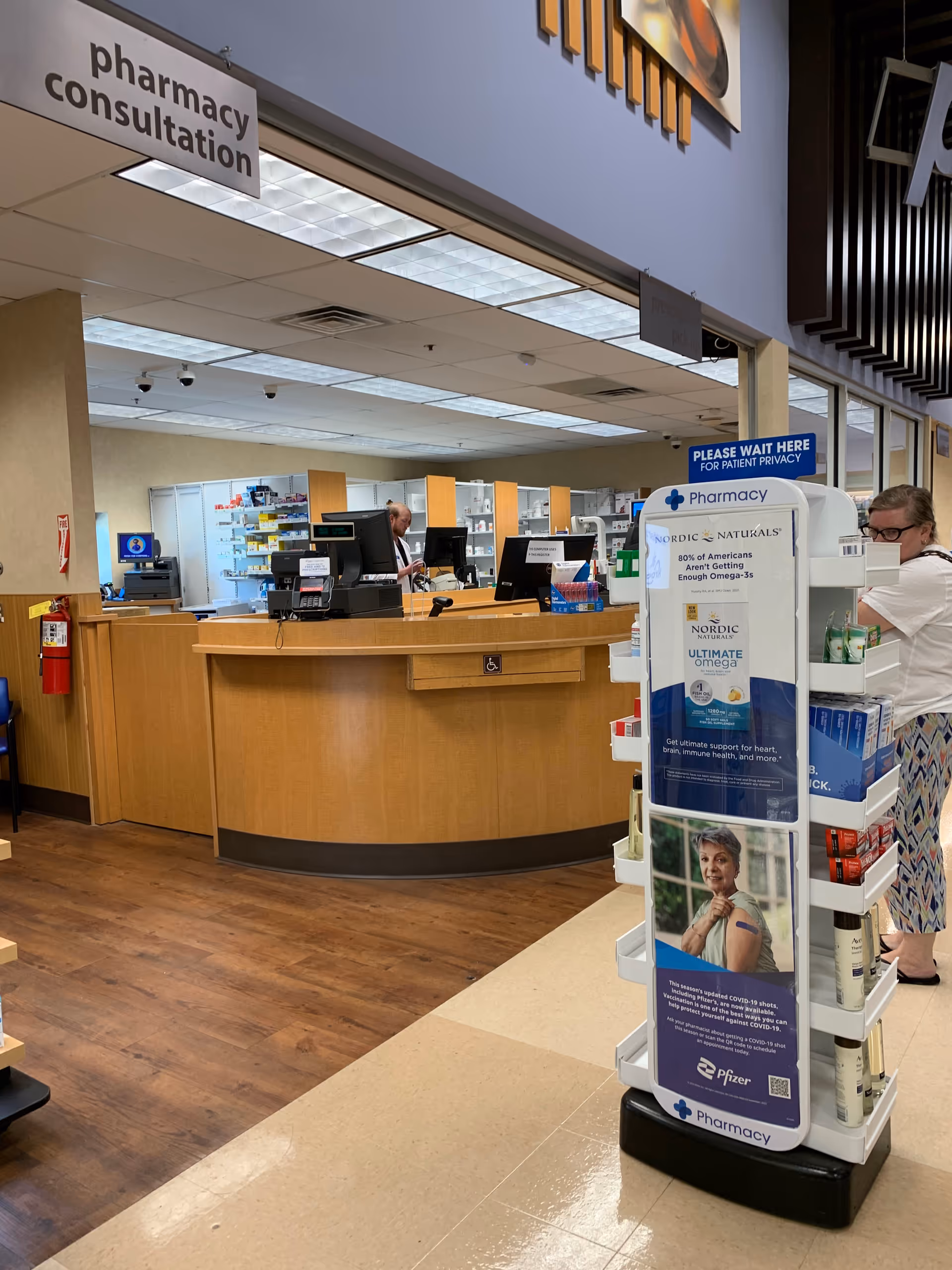 Inside a pharmacy area with a curved wooden counter, signage, and product display stand.