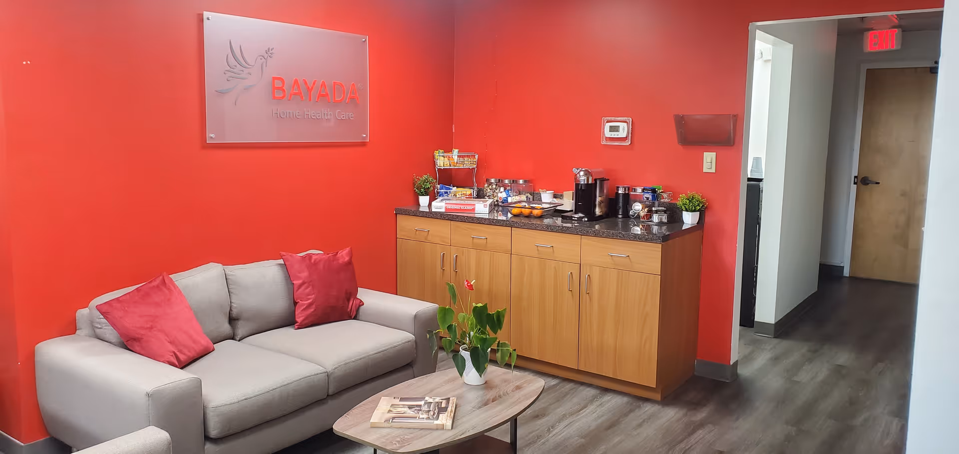 Red-walled BAYADA Assistive Care lounge with beige sofa, coffee table, and beverage counter.