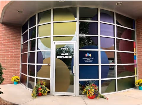 Curved glass façade with multicolored circular panels and a main entrance door flanked by flowers.