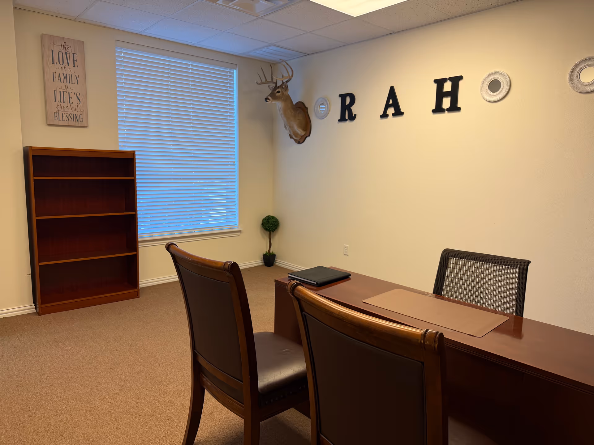 Office-style conference room with wooden desk, chairs, bookshelf, and wall decor.