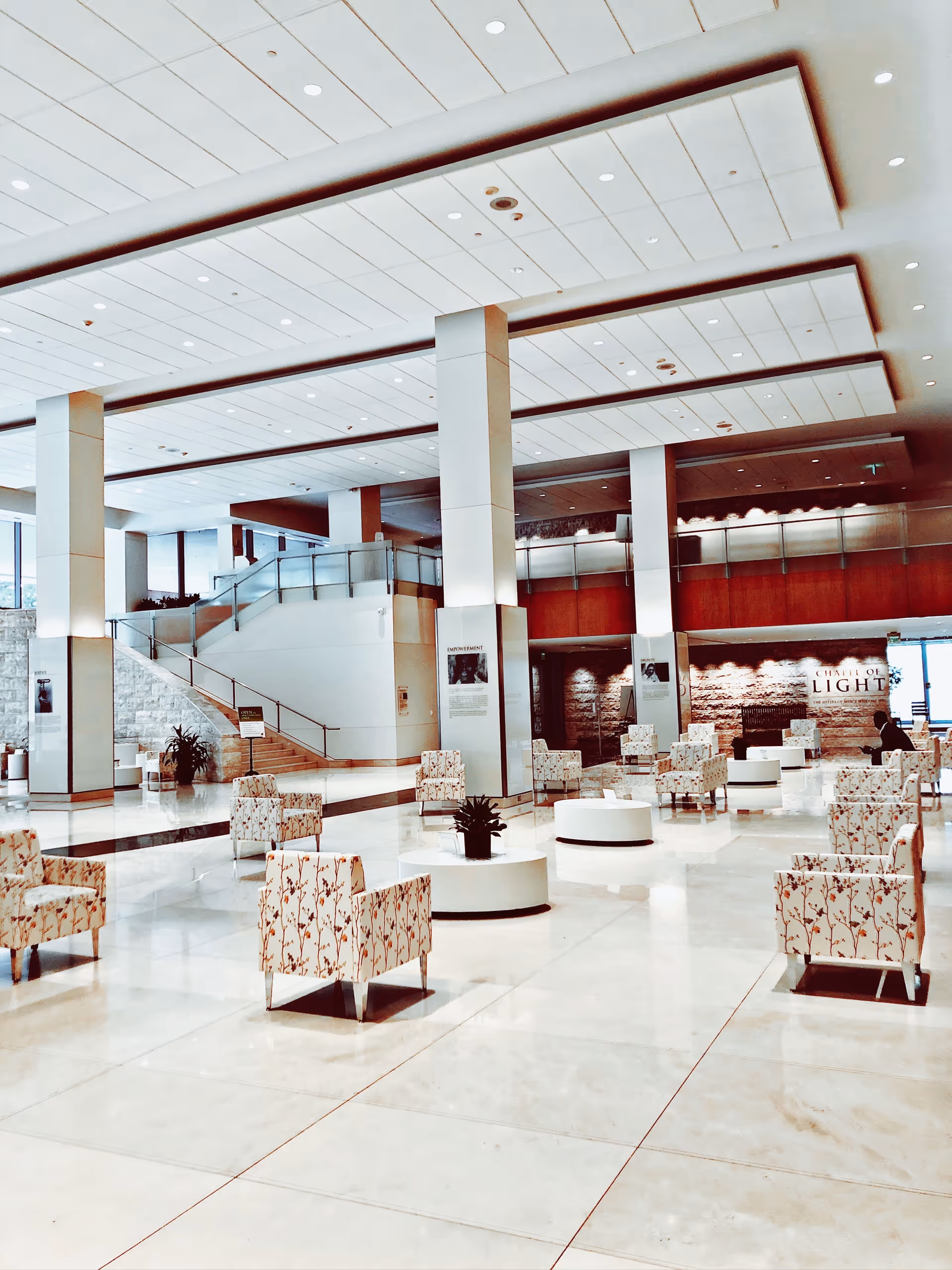 Bright hospital lobby with white tiled floor, pillars, stairs, and scattered patterned chairs.