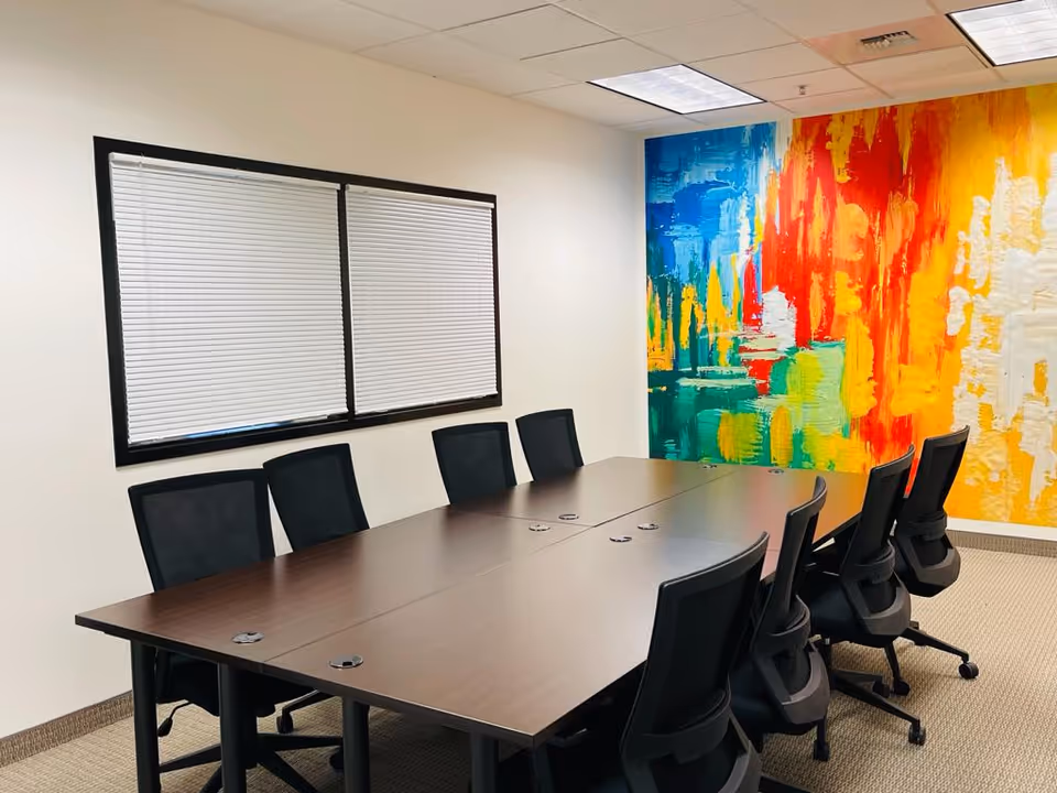 A long conference table with black chairs in a modern meeting room with a colorful mural.