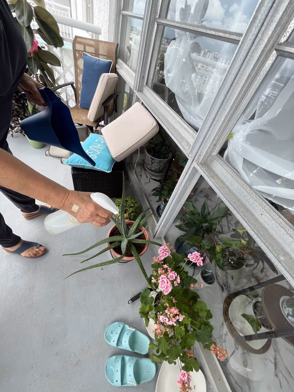 A person waters potted plants on a balcony with chairs and cushions.