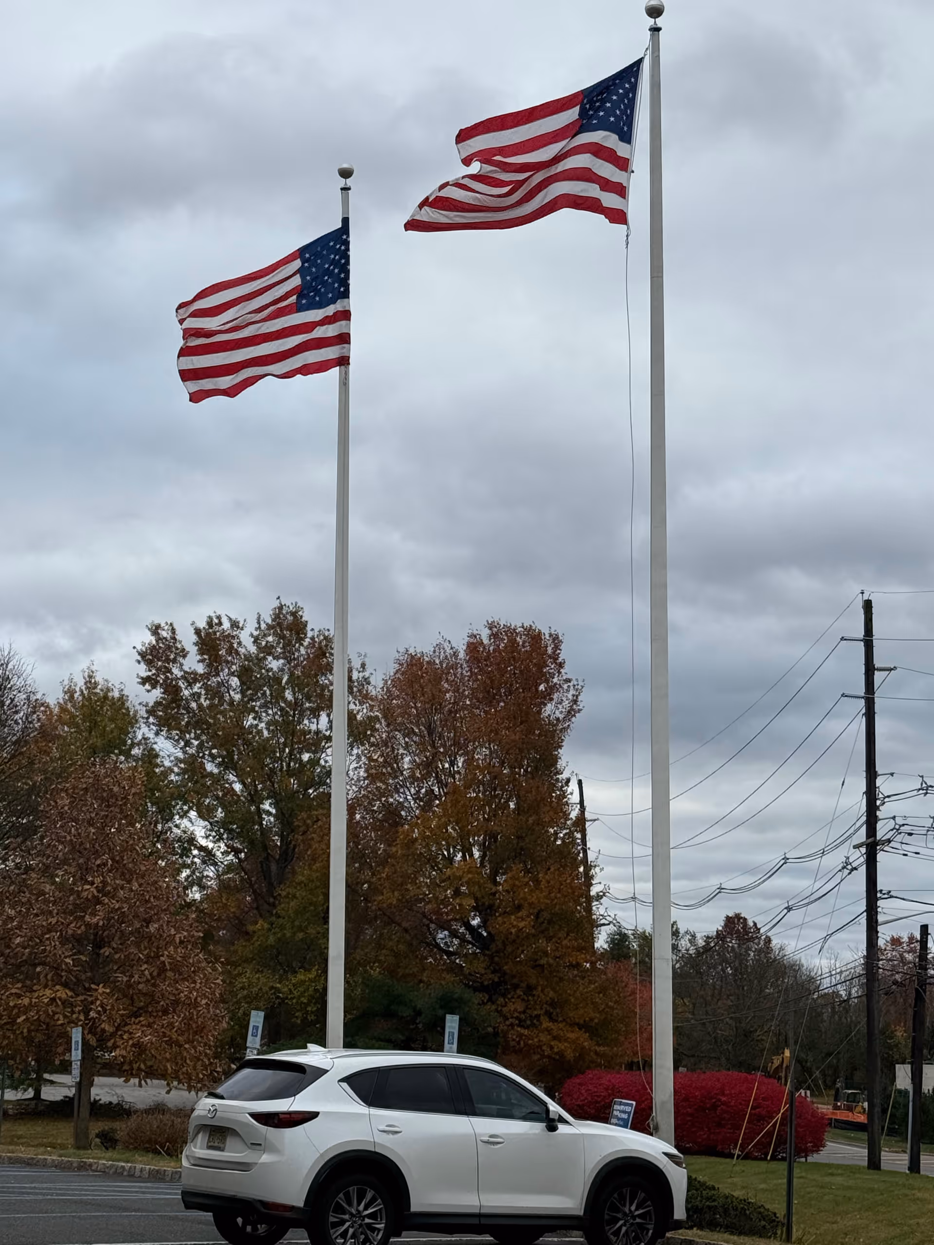 Two American flags on tall poles in a parking lot beside autumn trees.