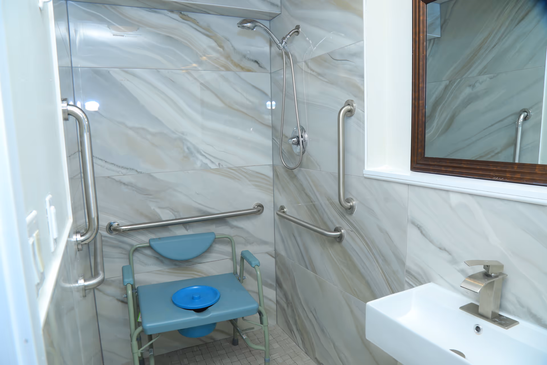Accessible bathroom with grab bars, blue commode chair, sink, and marble-tiled walls.