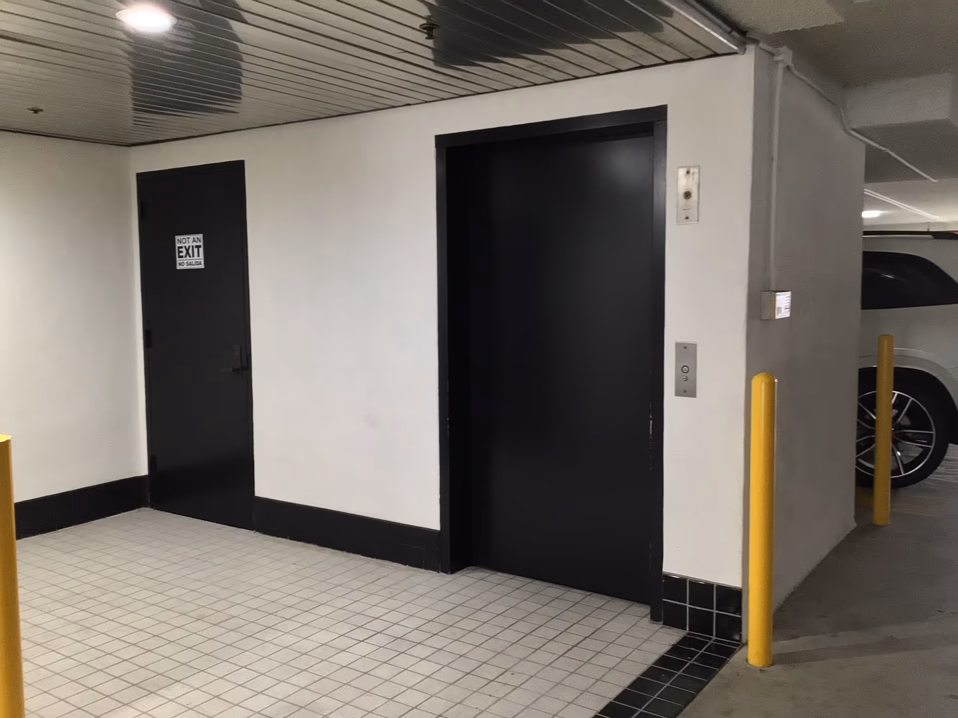 Underground parking area with elevator doors, a Not an Exit sign, tiled floor and yellow posts.