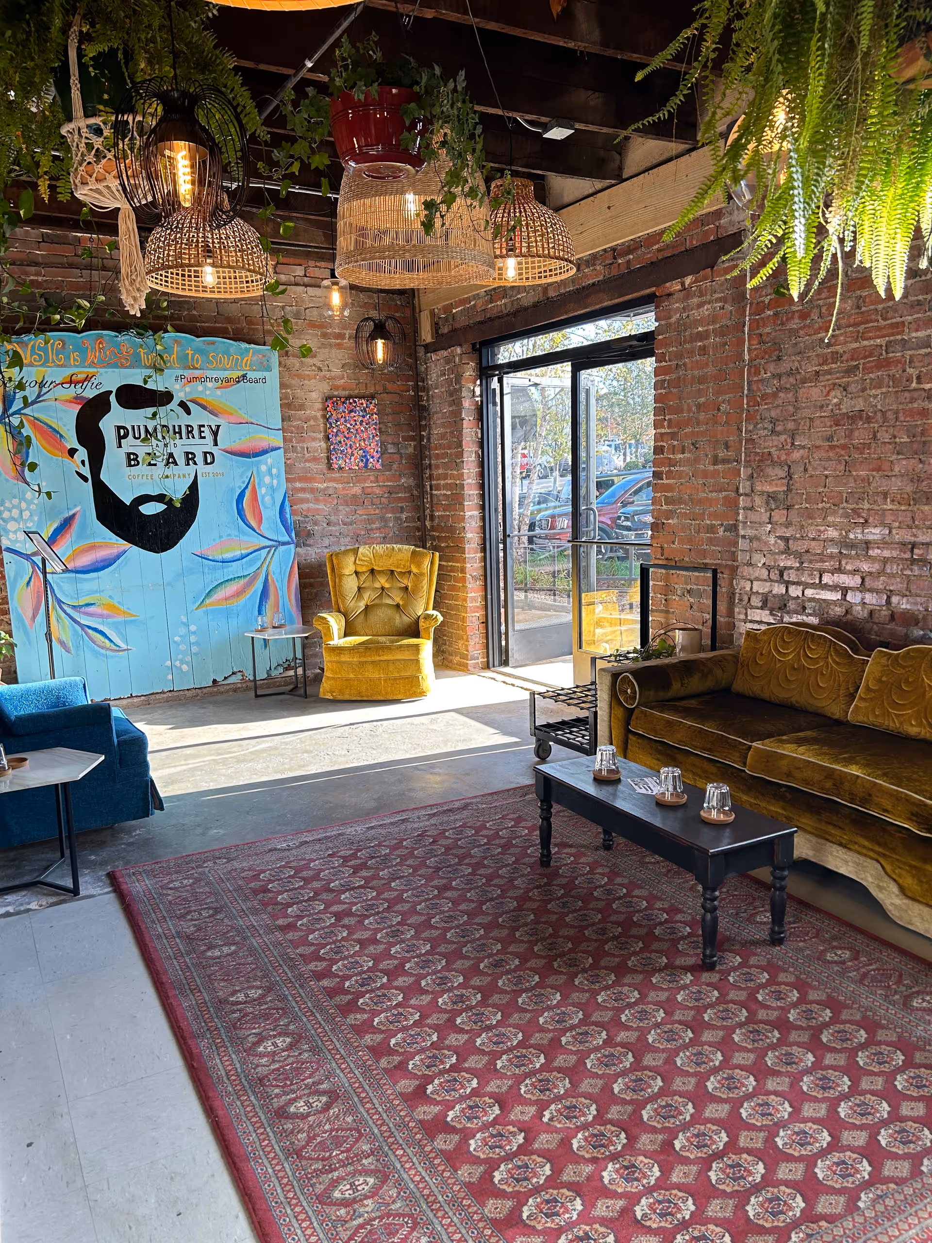 Industrial-style lounge with brick walls, hanging plants, colorful chairs, and a red patterned rug.