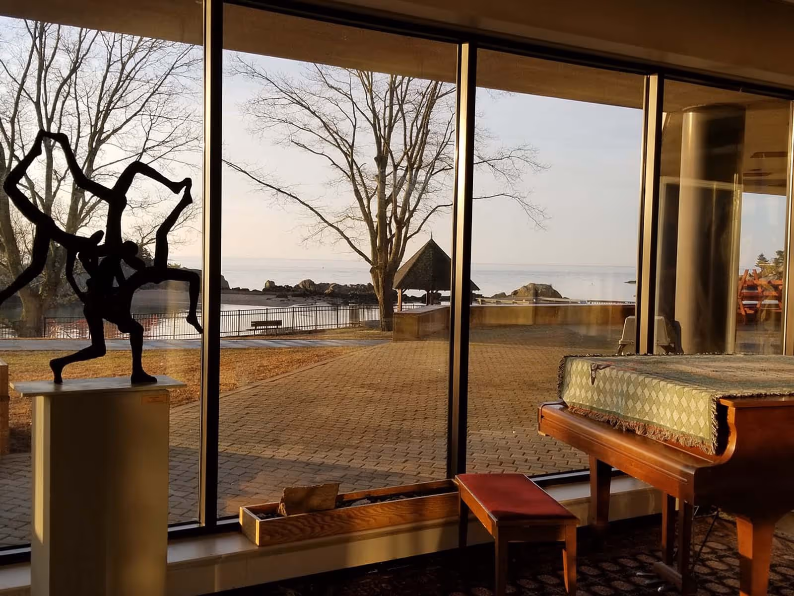 Sunlit hospice lounge with a grand piano, modern sculpture, and large windows overlooking water.
