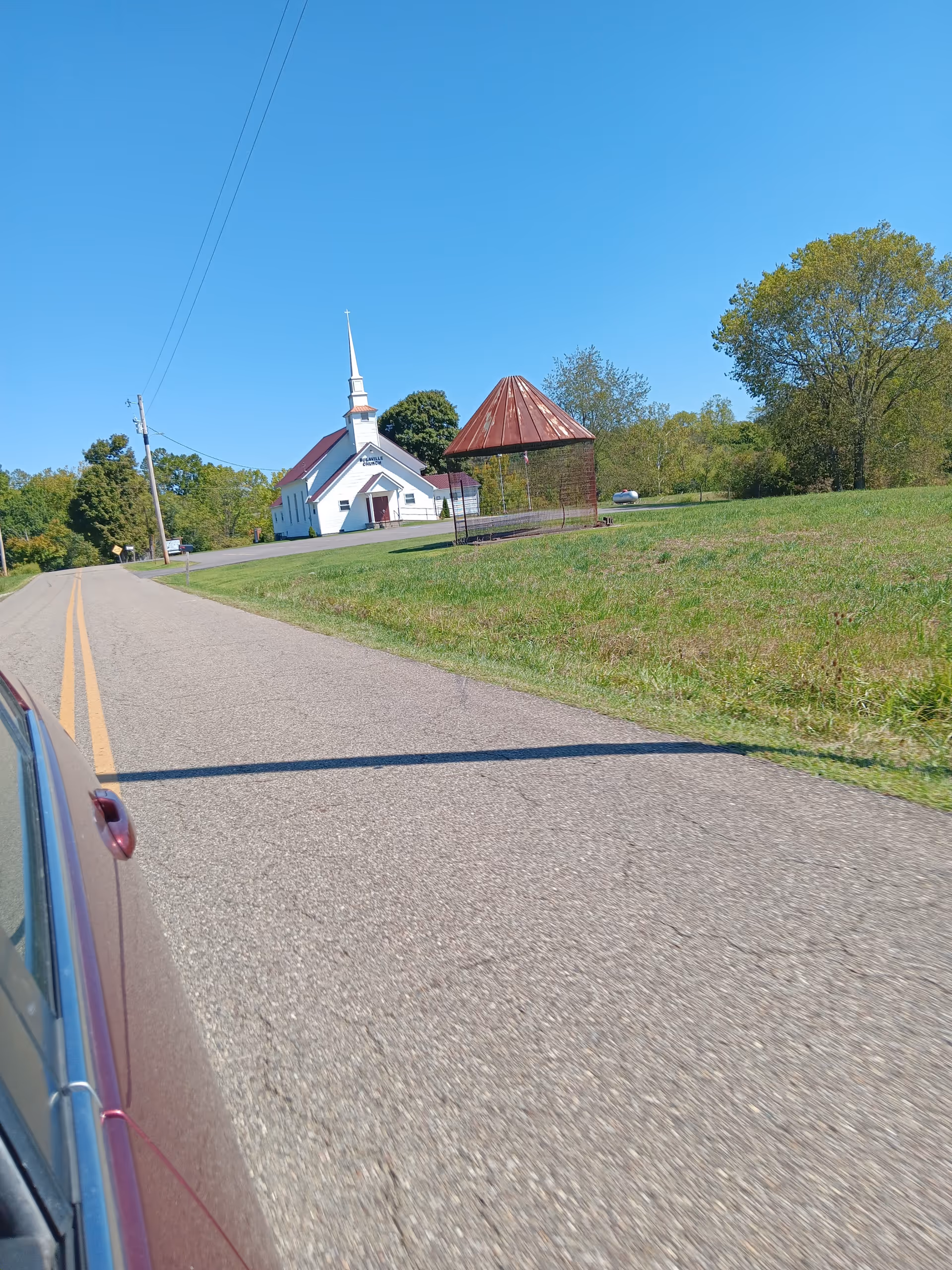 Roadside view of a white church with steeple and a metal gazebo on grassy land.