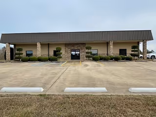 Front view of a single-story facility with stone columns, trimmed bushes, and a large parking area.