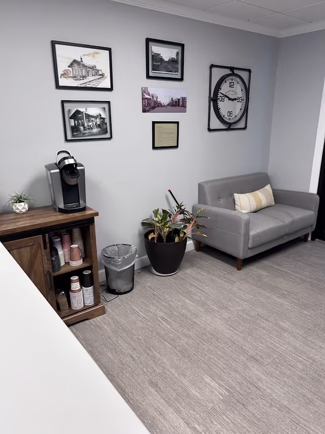 A small lounge area with a gray sofa, coffee station, plants, and wall art.